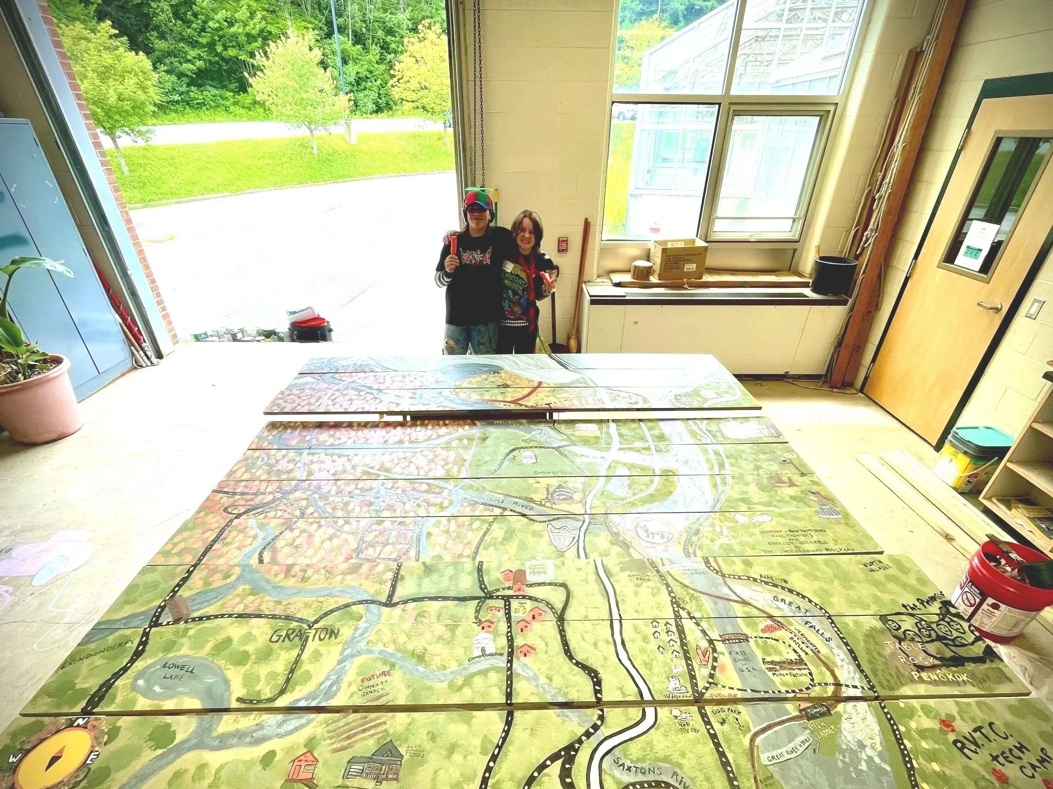 Two children standing inside a room near large windows, holding colorful items, with a large illustrated map on the table in front of them and greenery outside.