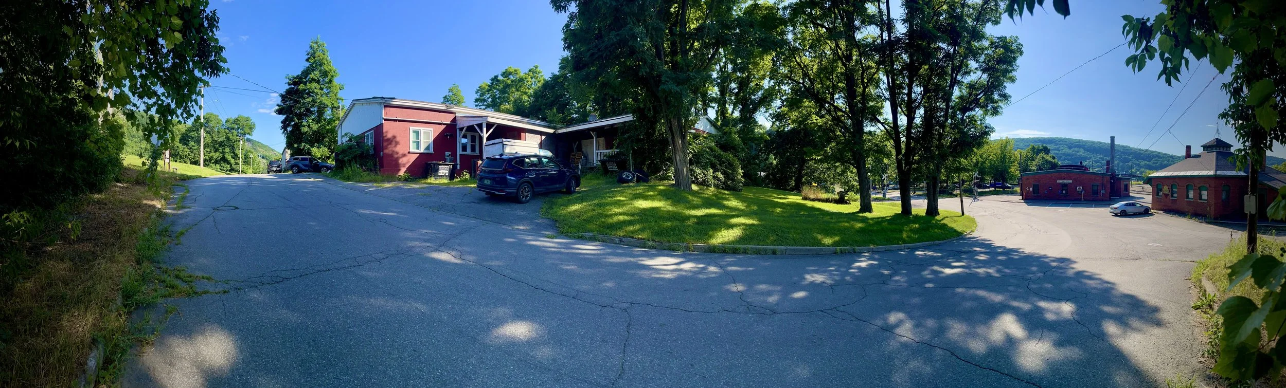 Panoramic view of a small town street with trees, cars parked along the road, a red house on the left, and a brick building on the right, under a bright blue sky.