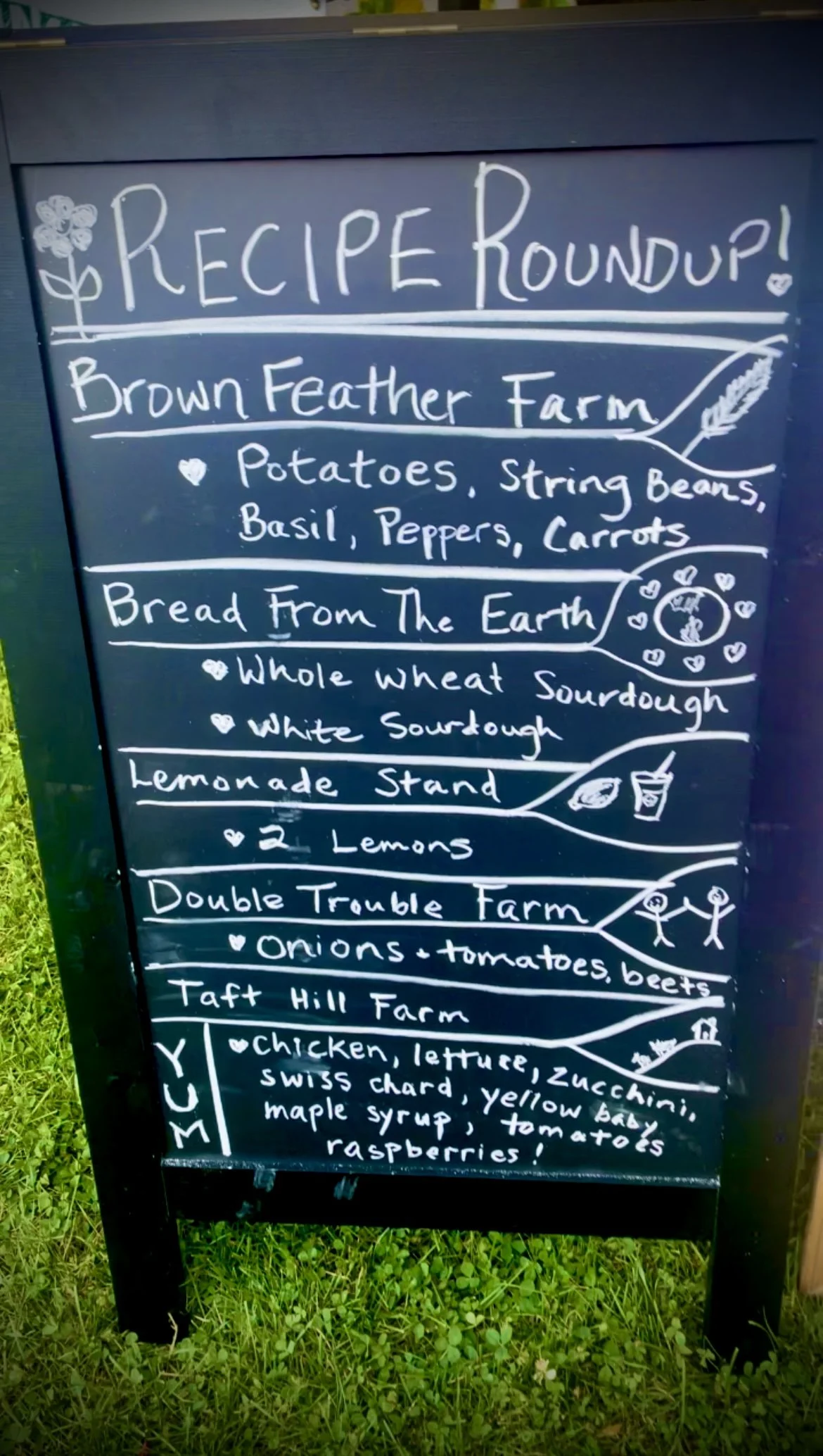 A black chalkboard sign with white handwritten text outlining different farm recipes and ingredients, including potatoes, onions, tomatoes, and other vegetables, with small decorative drawings of a flower, a grain sheaf, a clock, and stick figures.