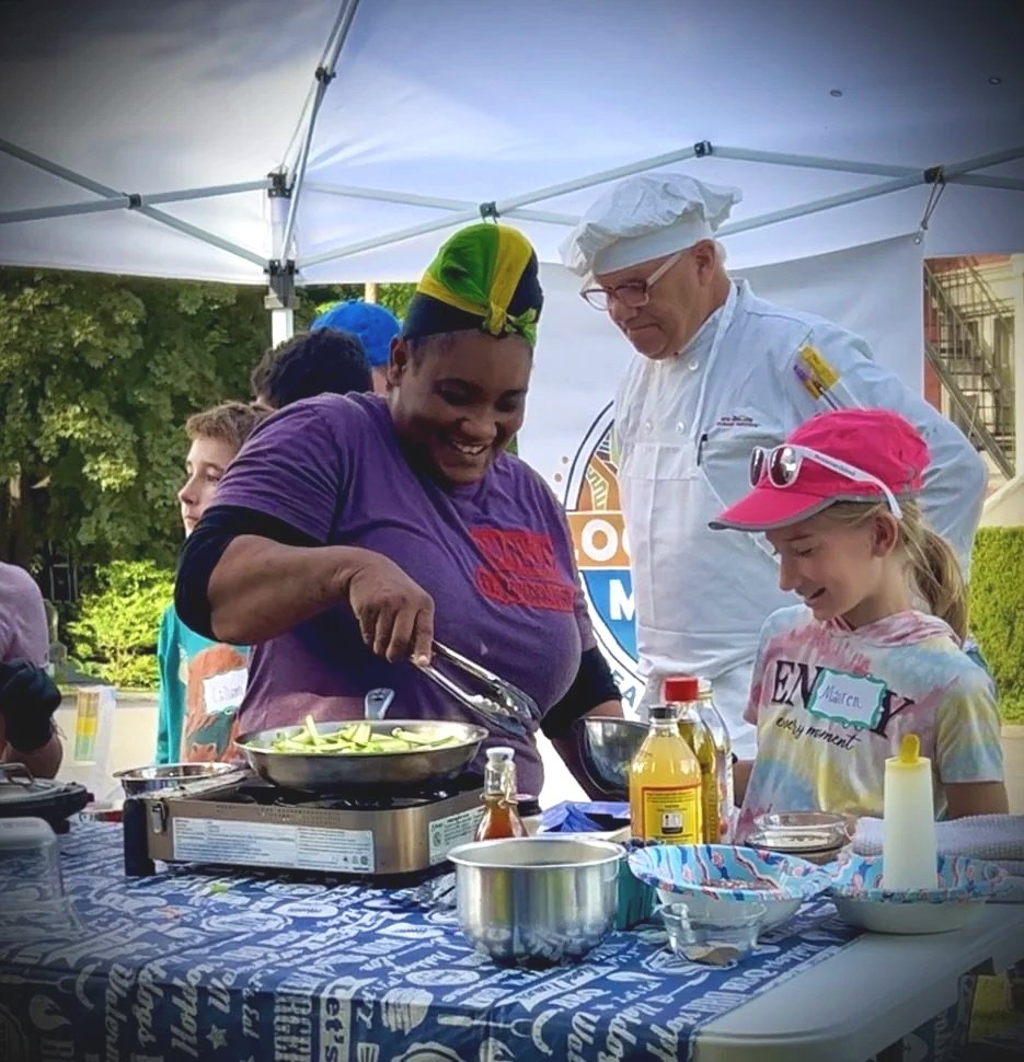 A woman wearing a purple shirt and a colorful headscarf cooks on a portable grill at an outdoor event. Several children and a chef in a white uniform and hat are watching her. The scene is under a white canopy trees and a building in the background.