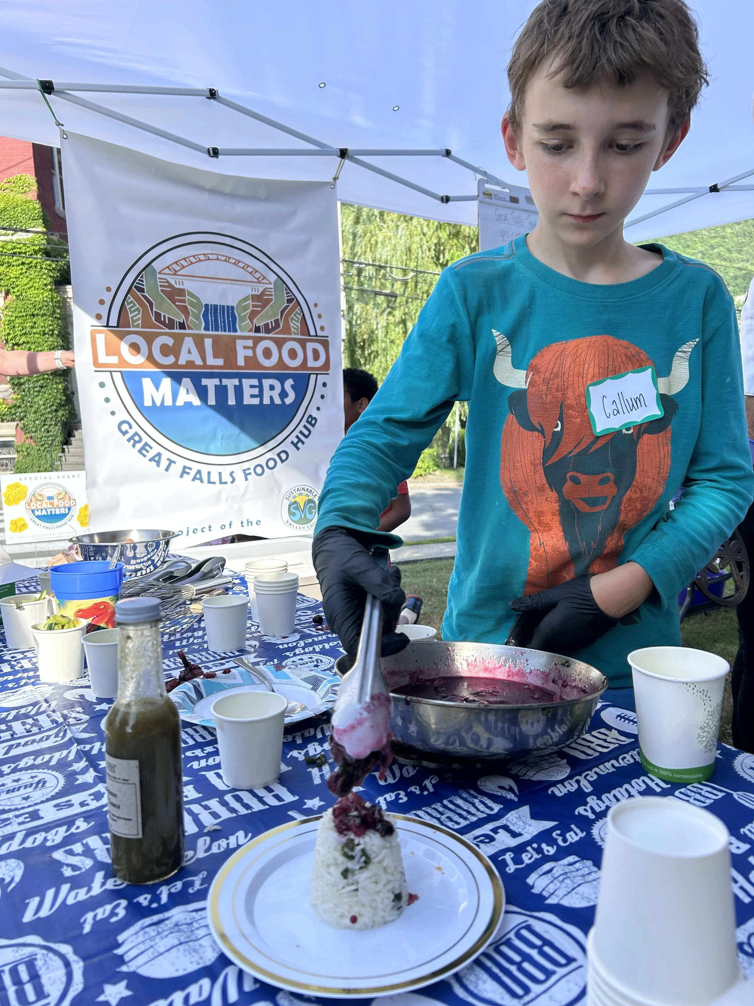 A young boy named Callum is serving berry sauce over a rice cake dessert with a Halloween-themed marshmallow topper at an outdoor event. There are cups, a bottle of syrup, and a sign that reads 'Local Food Matters' in the background.