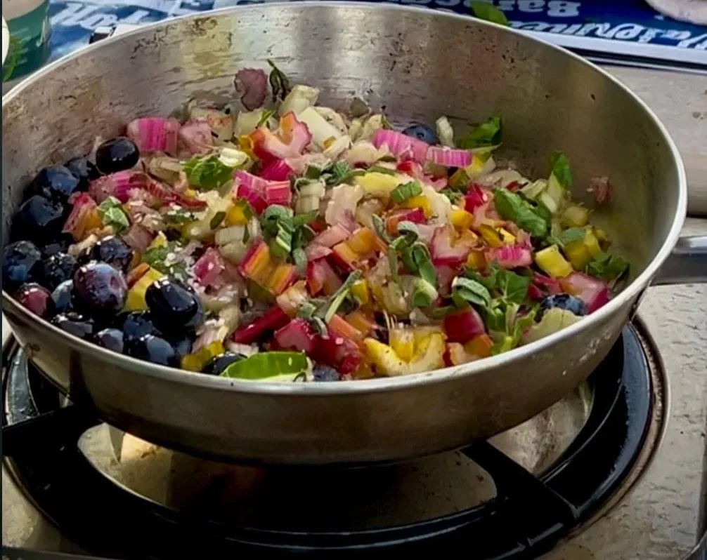 A frying pan containing a mixture of chopped vegetables, including blueberries, green onions, and rainbow chard, on a stove.
