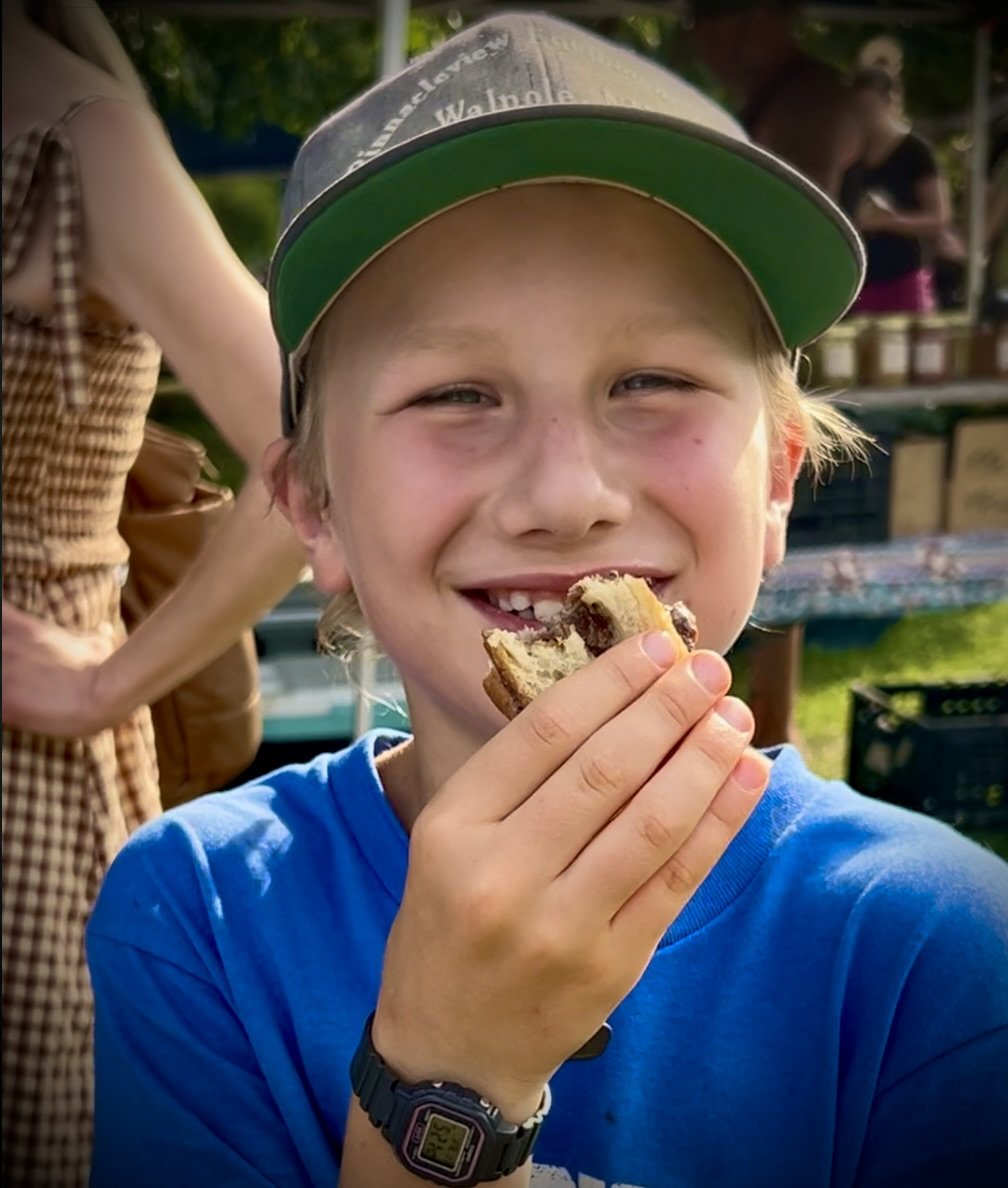 A young boy wearing a gray cap with green brim and a blue shirt, smiling and holding a chocolate chip cookie with bite marks near his mouth. Outdoor setting with market stalls and people in the background.
