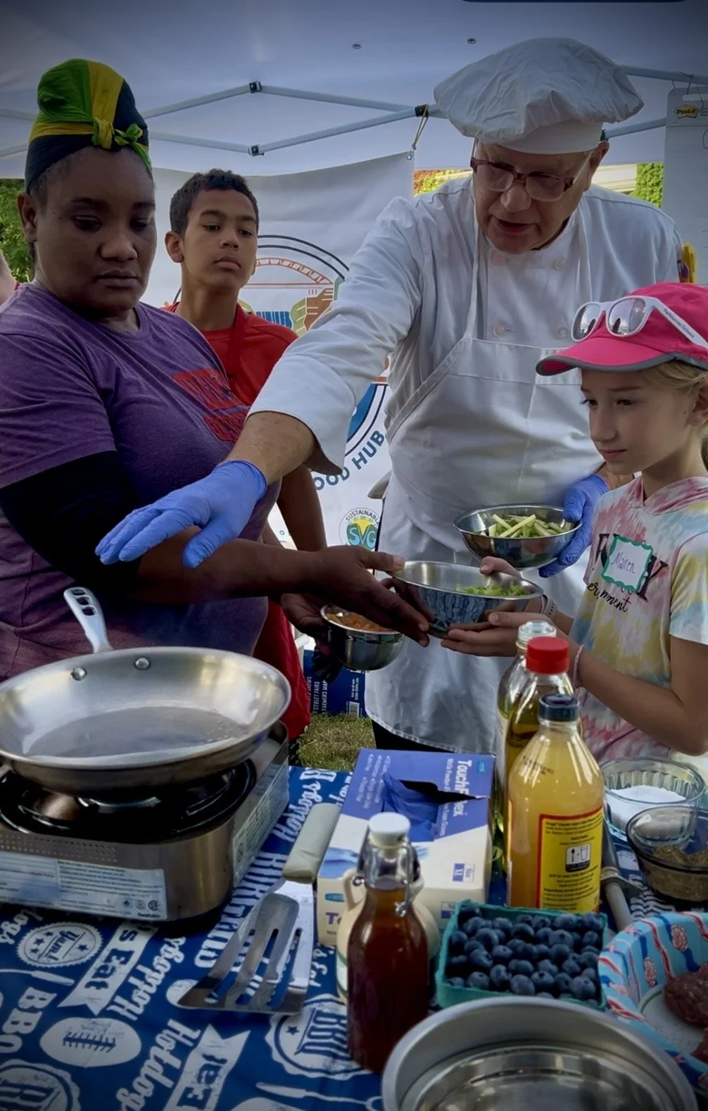 A chef in a white outfit and hat serves food to people at a food stand, with children and adults waiting and receiving bowls of food outdoors.