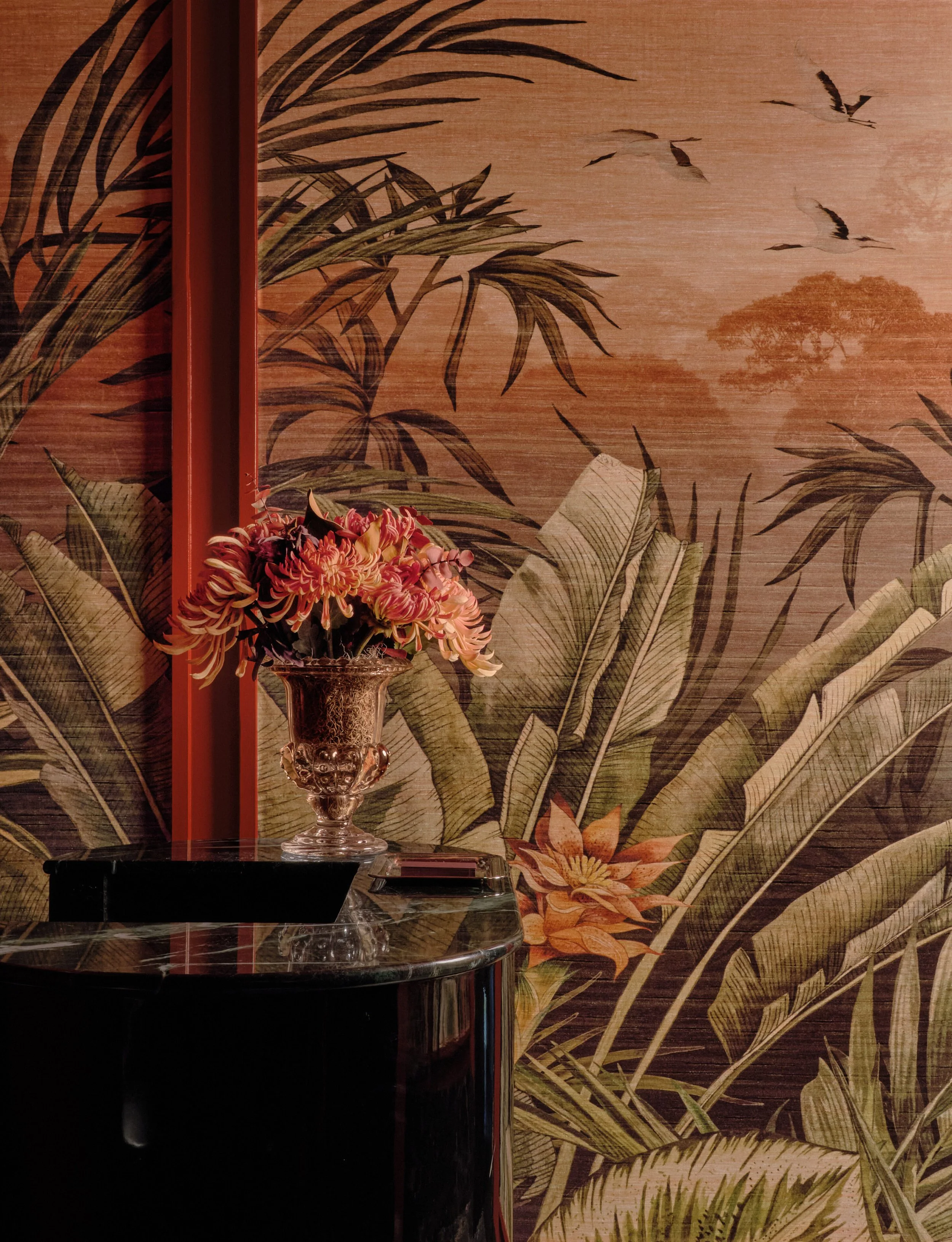 Bowie Flowers. A decorative wall with tropical leaves and birds, featuring a red vertical divider and a floral bouquet in a crystal vase on a black marble table.