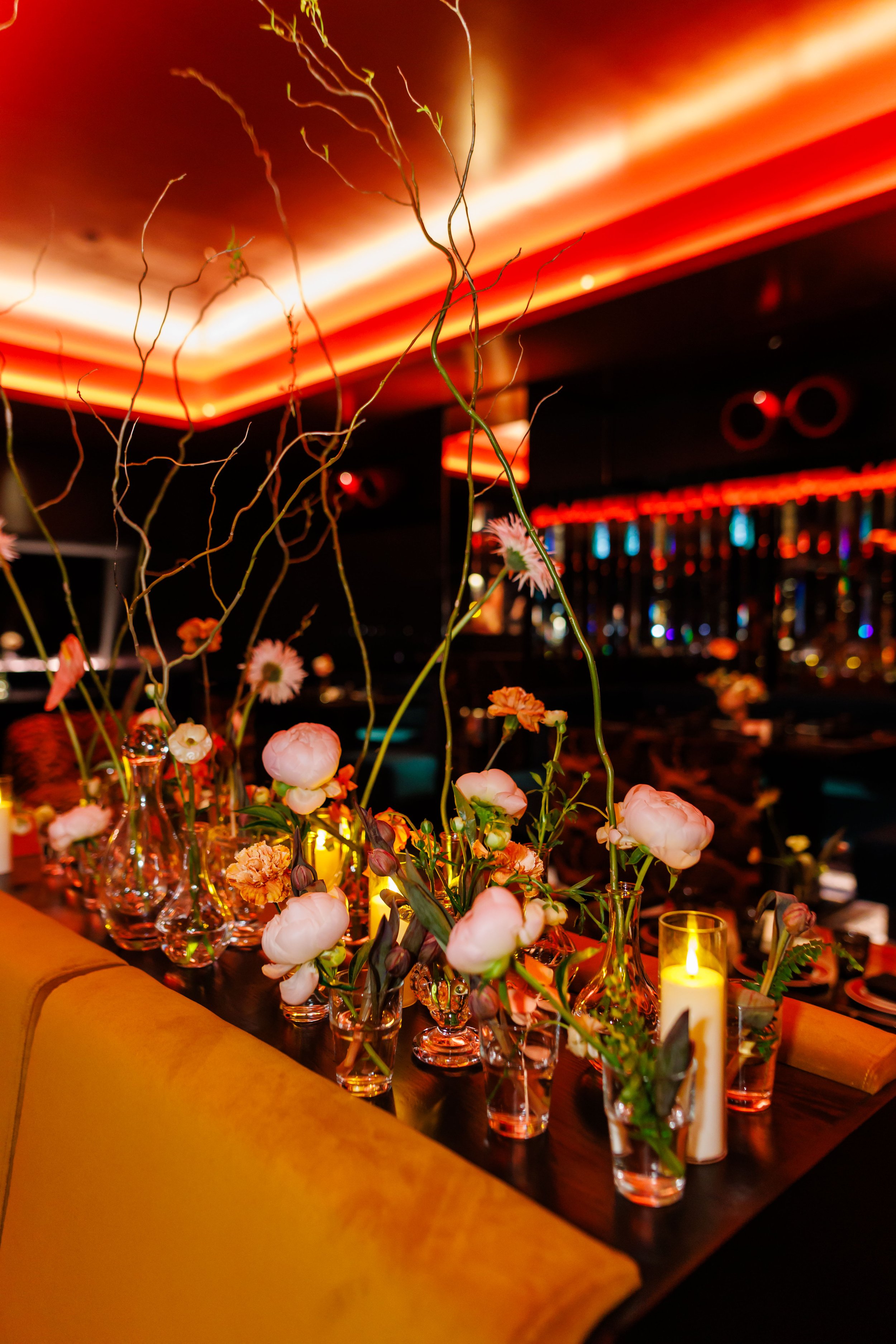 Veronica Beard event at Bowie, table decorated with various flowers in glass vases and candles, set in a dimly lit venue with red and orange lighting and a bar in the background.