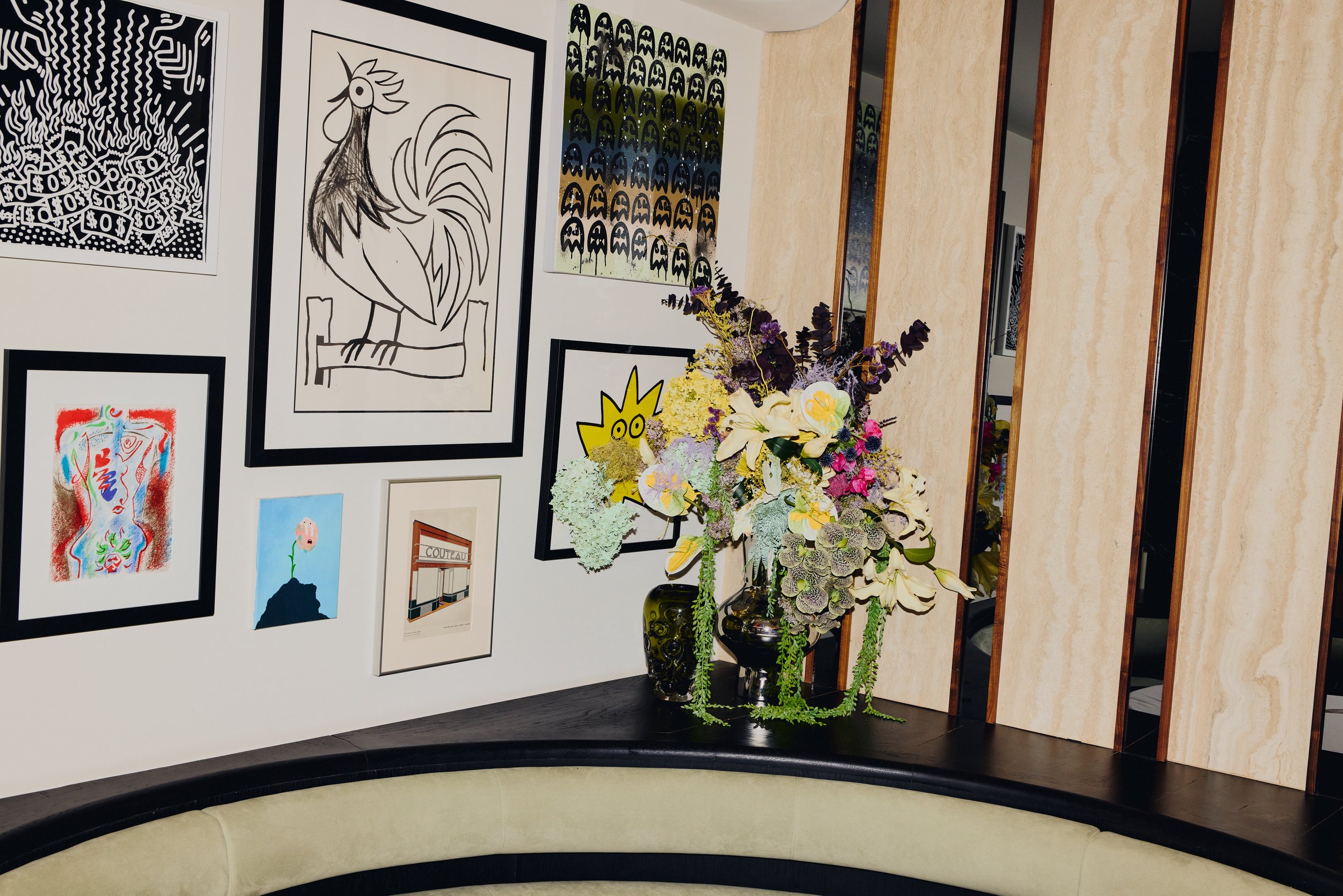 Dorsia Flowers. Gallery wall with various artworks, including a line drawing of a rooster, a colorful abstract piece, and text-based art, with a large floral arrangement in front and mirrored doors reflecting some of the art pieces.