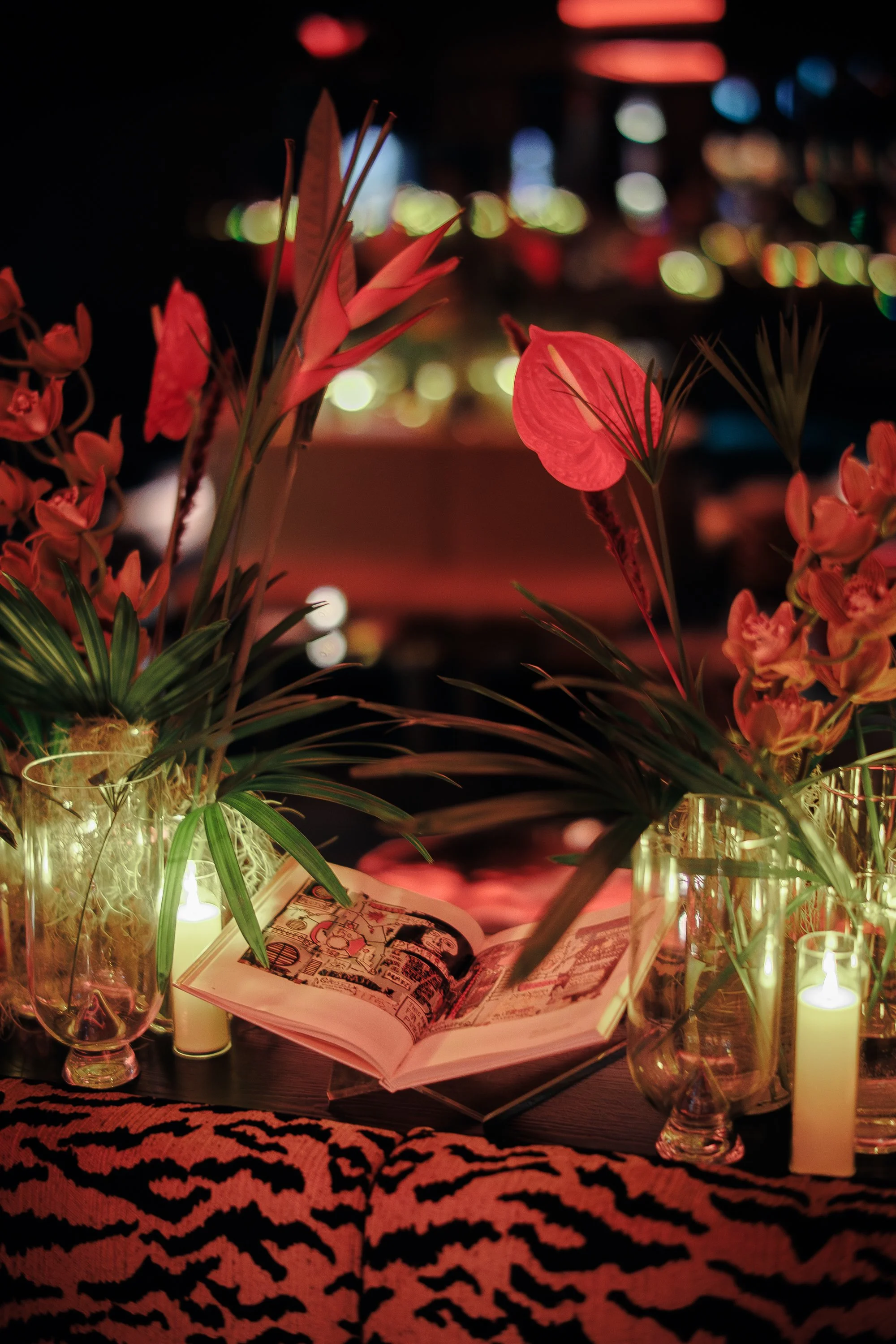 SOHO House event at Bowie. Cities without Houses, Montreal. Dimly lit lounge area with a plush, red and black patterned sofa, decorated with tall floral arrangements illuminated by candles, and ambient ceiling lighting.