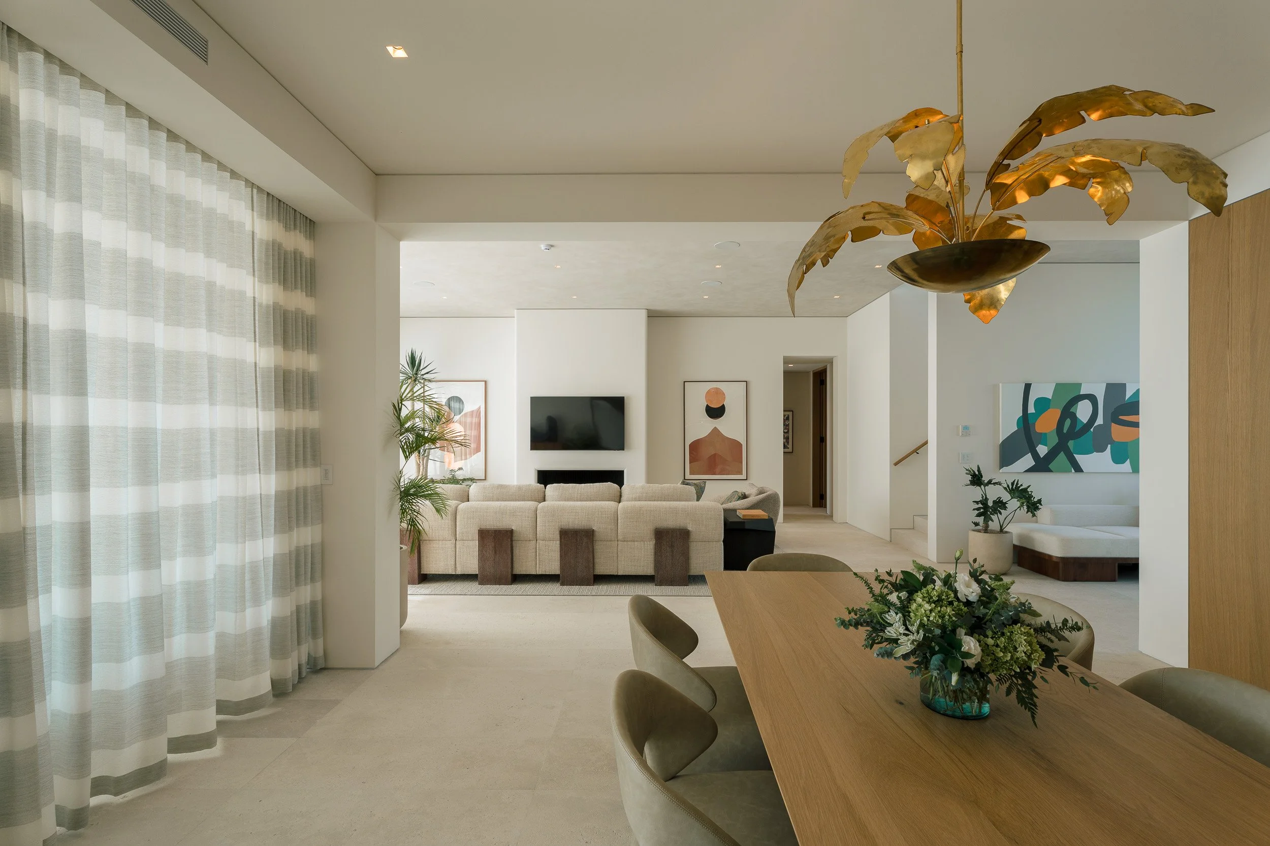 Interior of a modern living and dining area with a beige couch, abstract art on walls, a large window with striped curtains, and a wooden dining table with a flower arrangement.