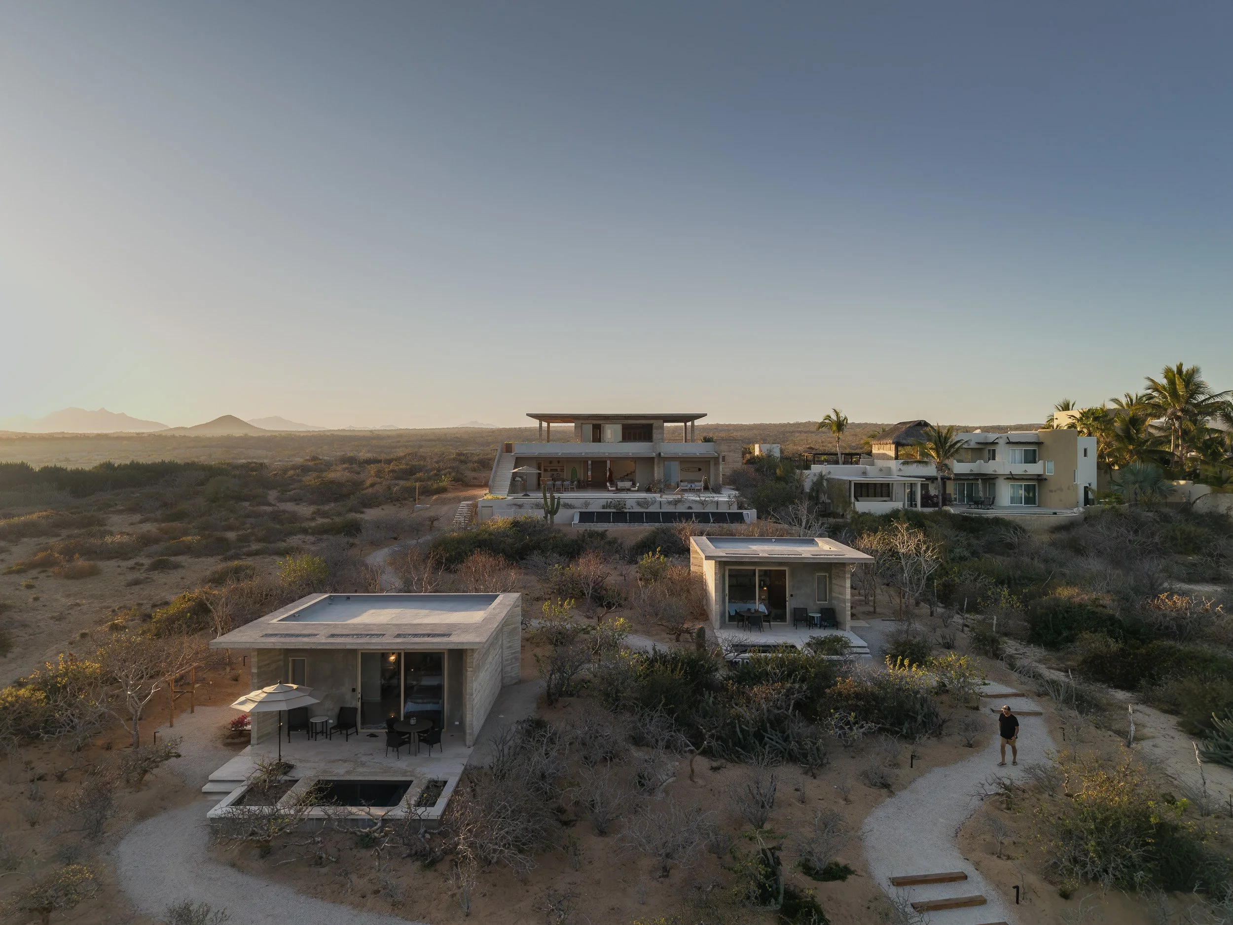 Aerial view of modern houses in a desert landscape at sunset, with mountains in the background and a man walking on a path.