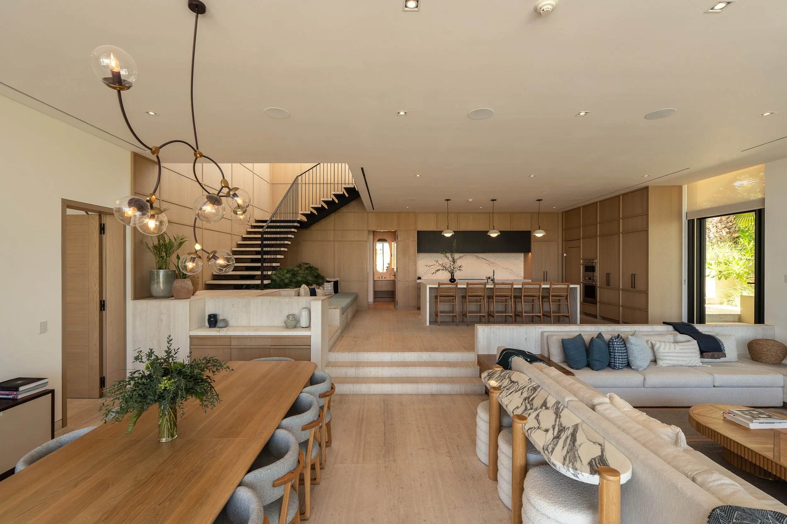 Open-concept living space with a dining table, modern chandelier, staircase, and kitchen area with an island and wooden cabinetry.