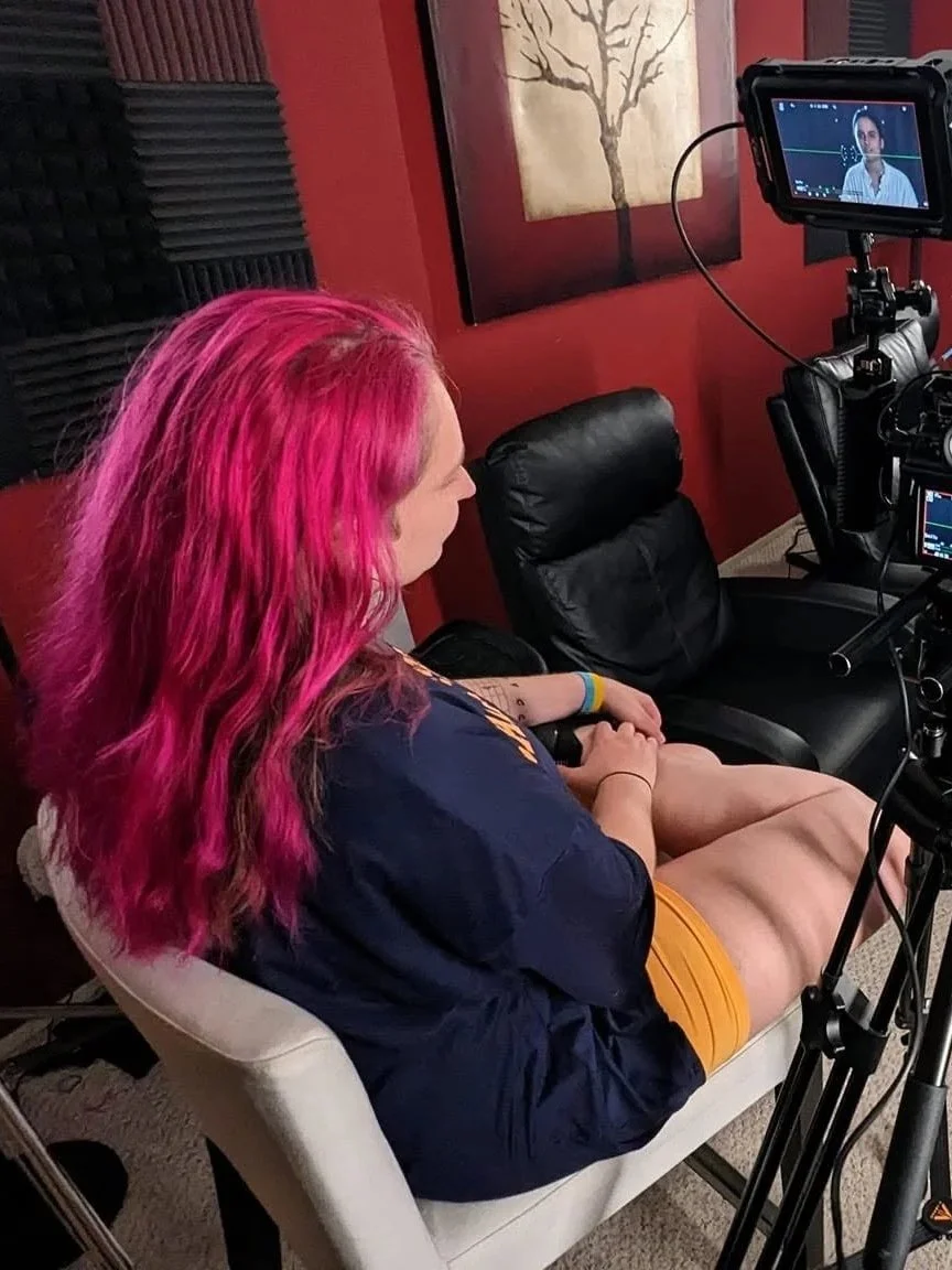 A woman with pink hair sitting on a chair in a recording studio, looking at a camera monitor.