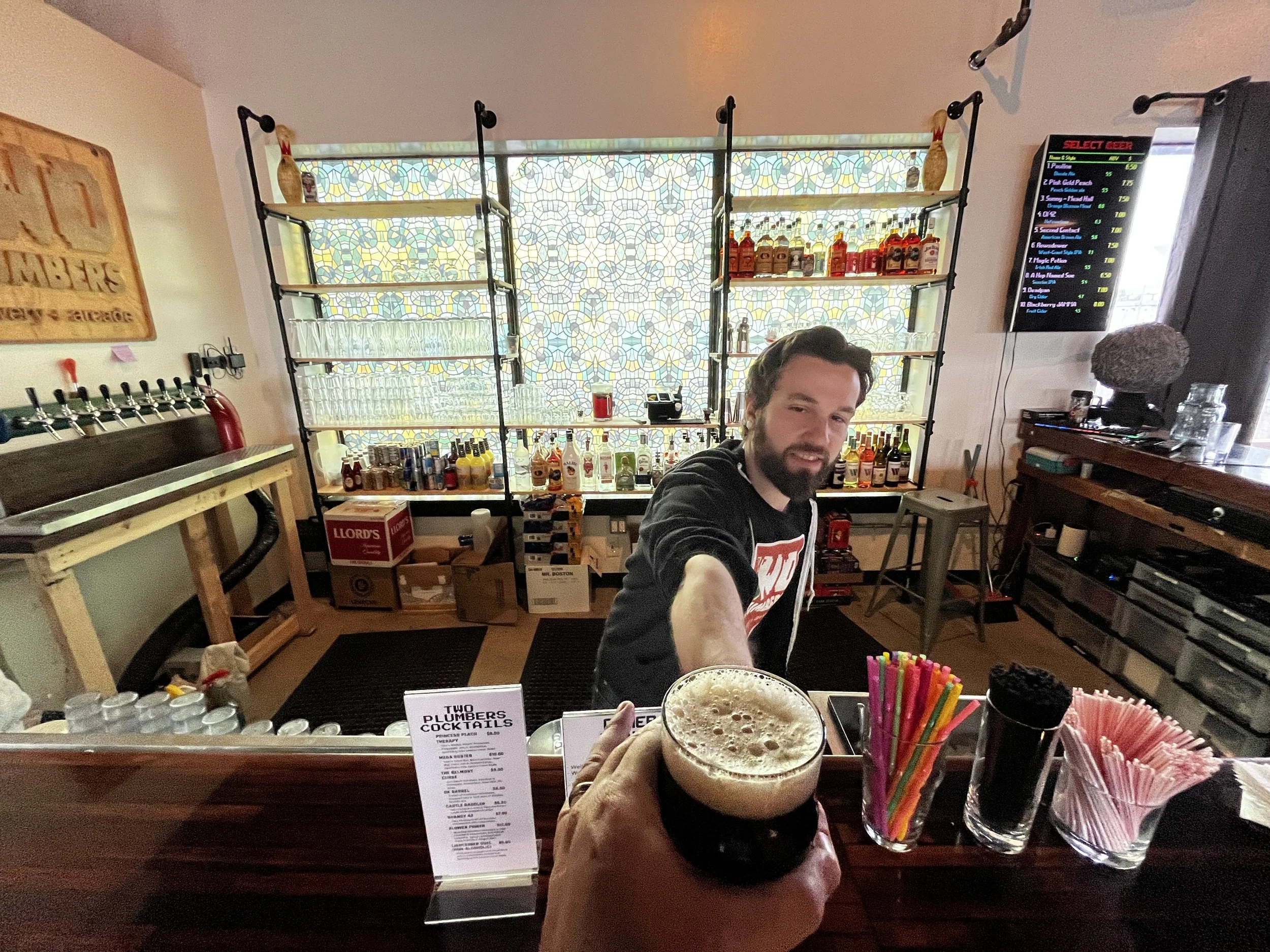 The image shows a bar scene with a man smiling. The foreground features a hand holding a frothy drink. Behind him, shelves stocked with liquor bottles are visible, and a man is partially captured in the reflection. To the right, a digital menu board displays drink options and prices. The decor includes a stained glass window and various bar tools and supplies.