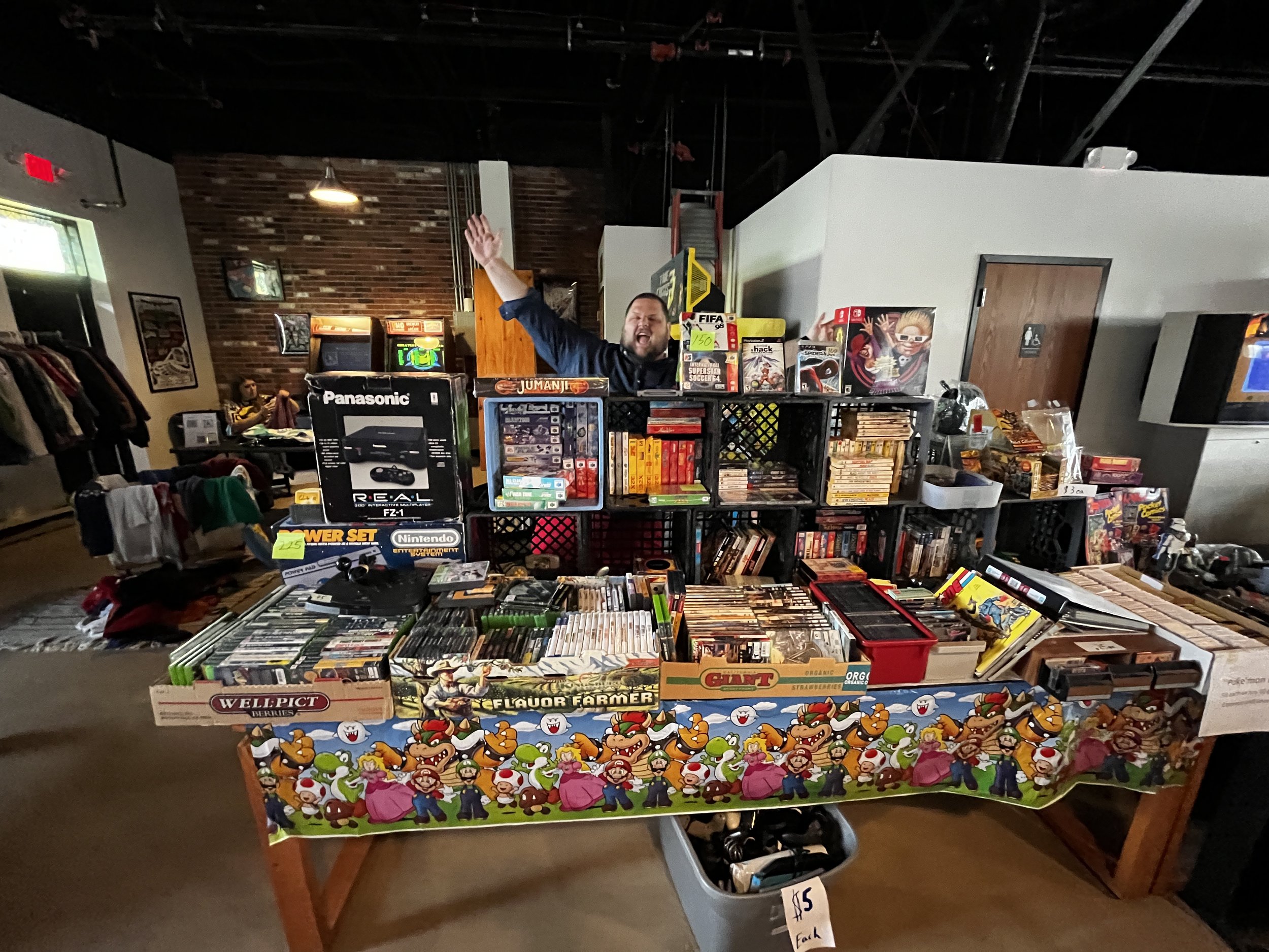 A man behind a table at a flea market, smiling and raising his hand. The table displays various video games, movies, and toys, including Nintendo Wii packages and Super Mario merchandise. There are racks of clothes and other shoppers in the background.