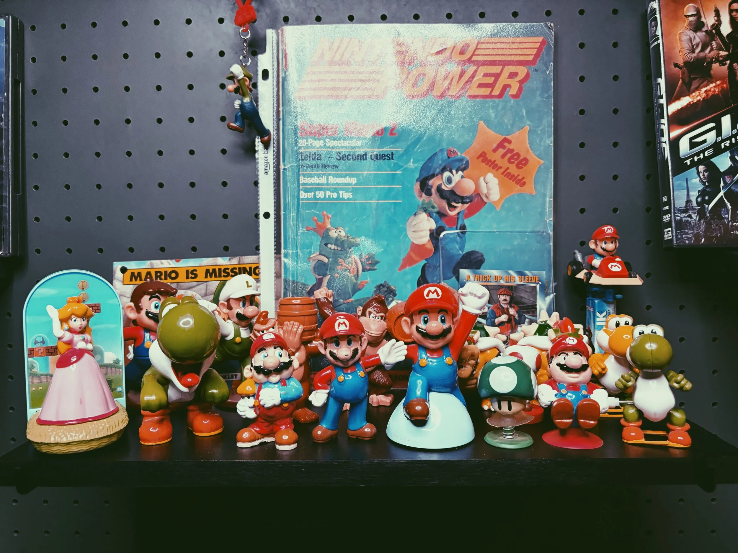 A collection of Super Mario characters and figurines on a black display shelf, including Mario, Luigi, Princess Peach, Yoshi, Toad, and others, surrounding a vintage Super Mario magazine.