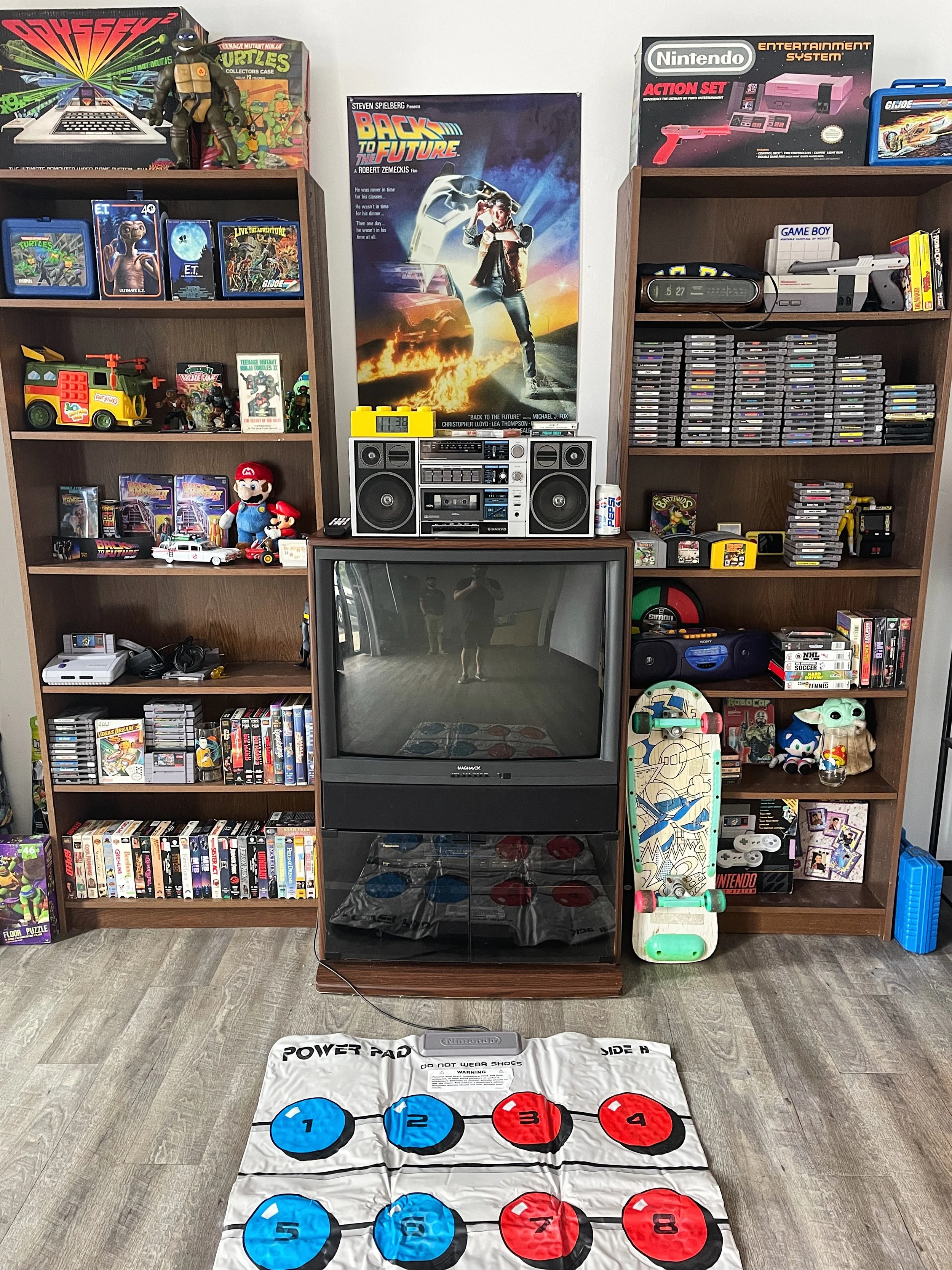 A room filled with vintage video game and pop culture collectibles, including shelves of video games and toys, a television, a Boombox, and a game mat on the floor with numbered circles in blue and red.