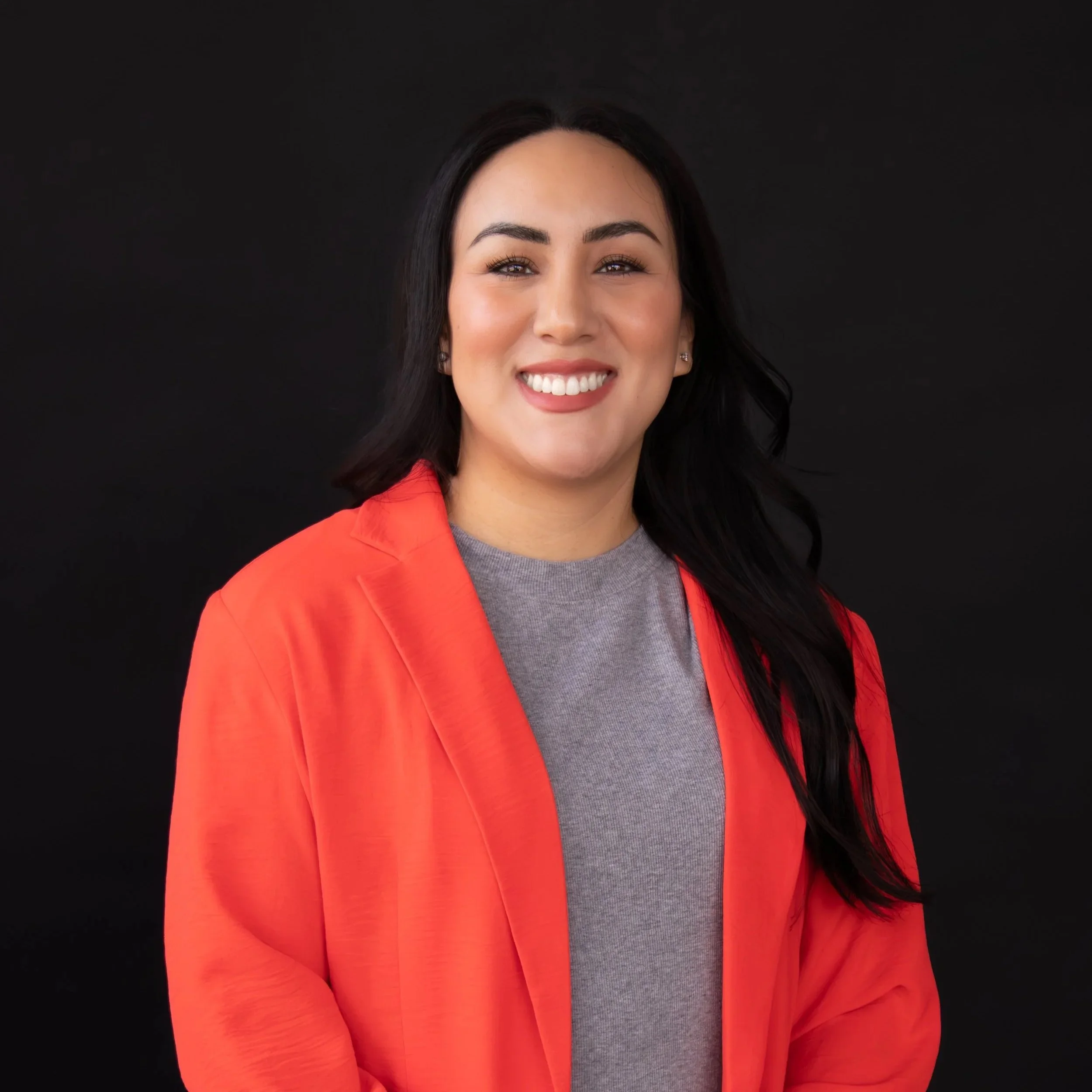 A woman with dark hair and earrings smiling, wearing a red jacket over a gray shirt, standing against a plain black background.