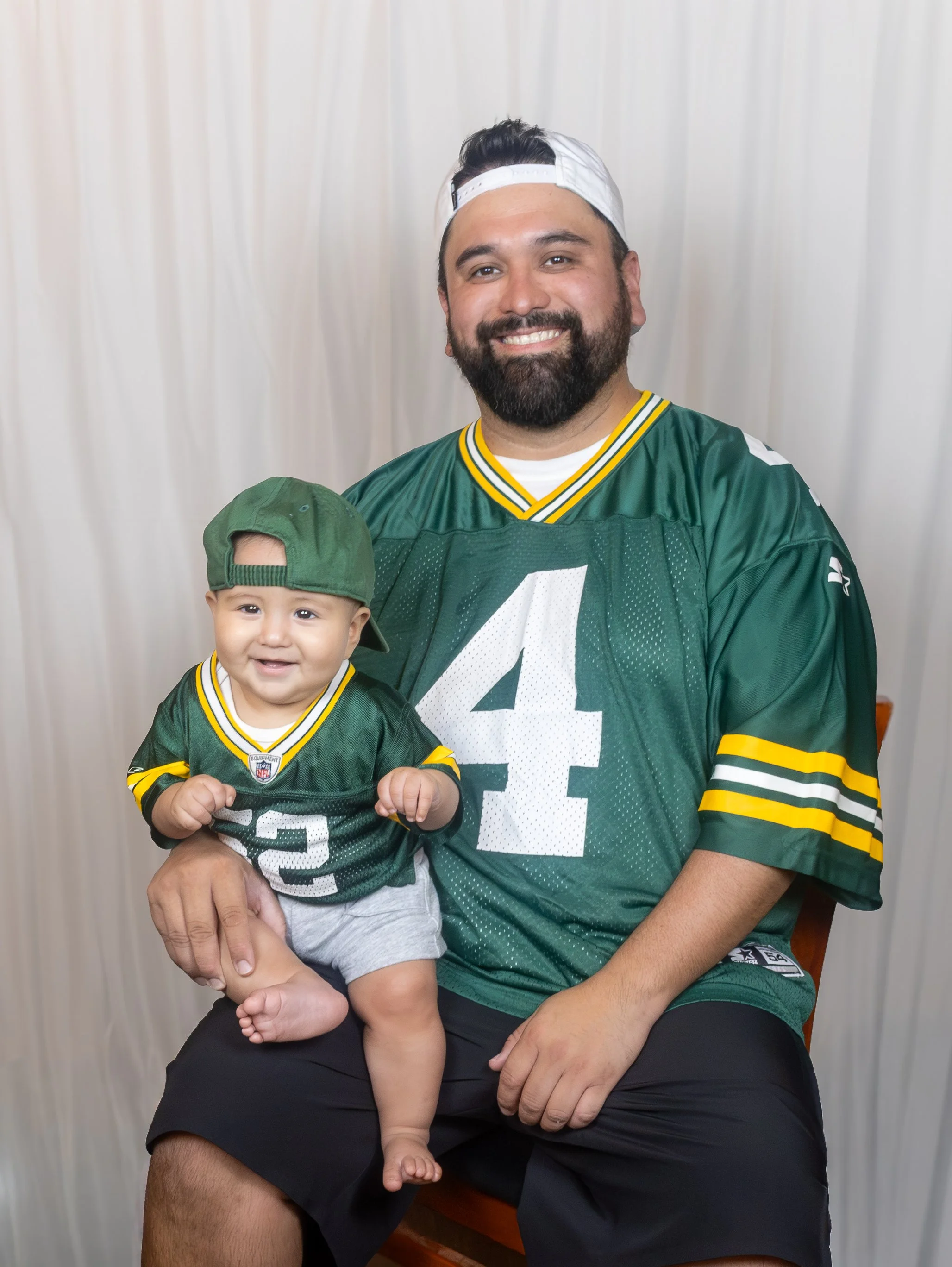A man with a beard and a white cap sitting on a wooden chair, smiling, holding a young boy with brown hair, a green cap, and a Green Bay Packers football jersey.