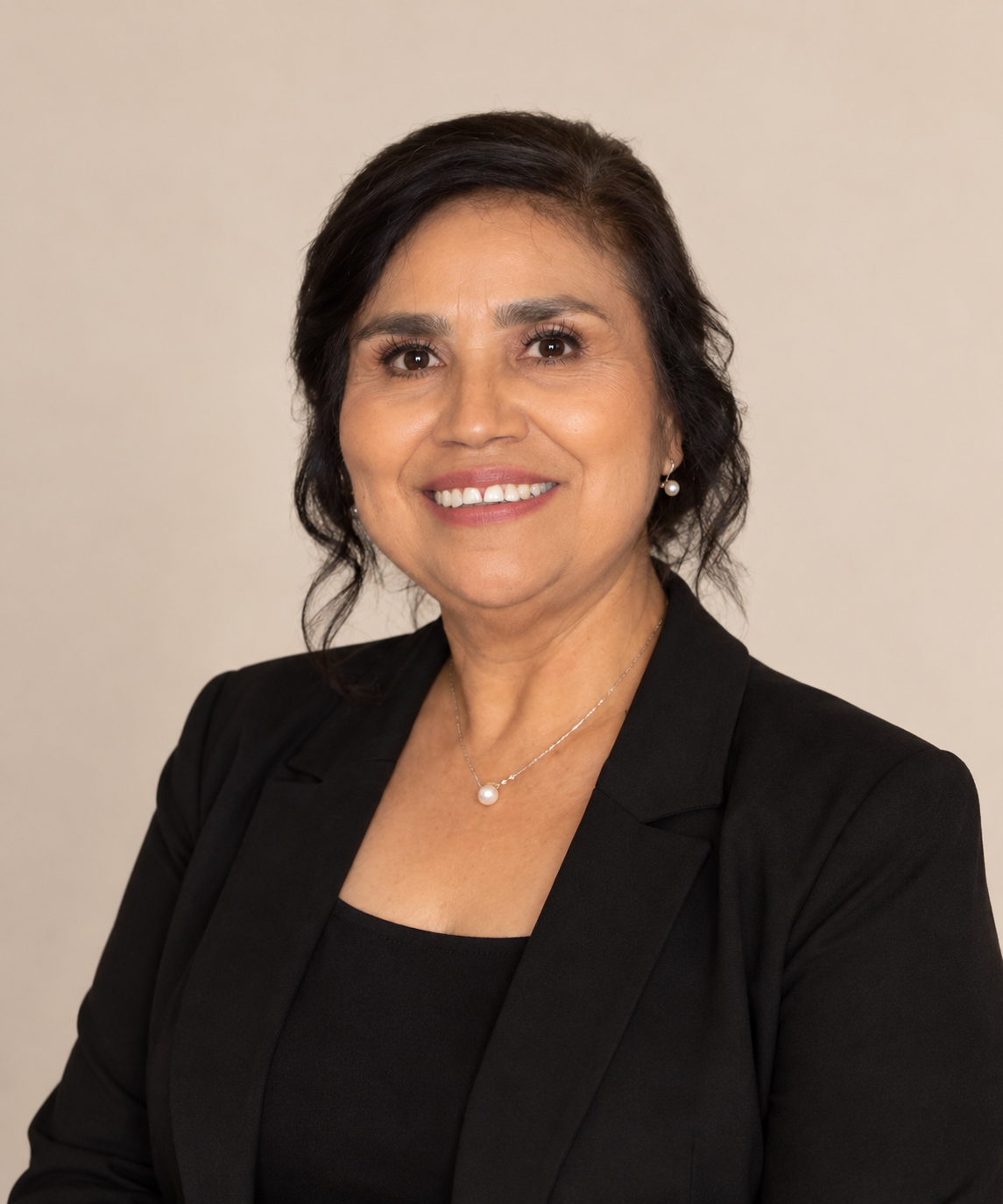 Professional headshot of a middle-aged woman with dark hair, wearing a black blazer, pearl earrings, and a pearl necklace, smiling against a plain beige background.