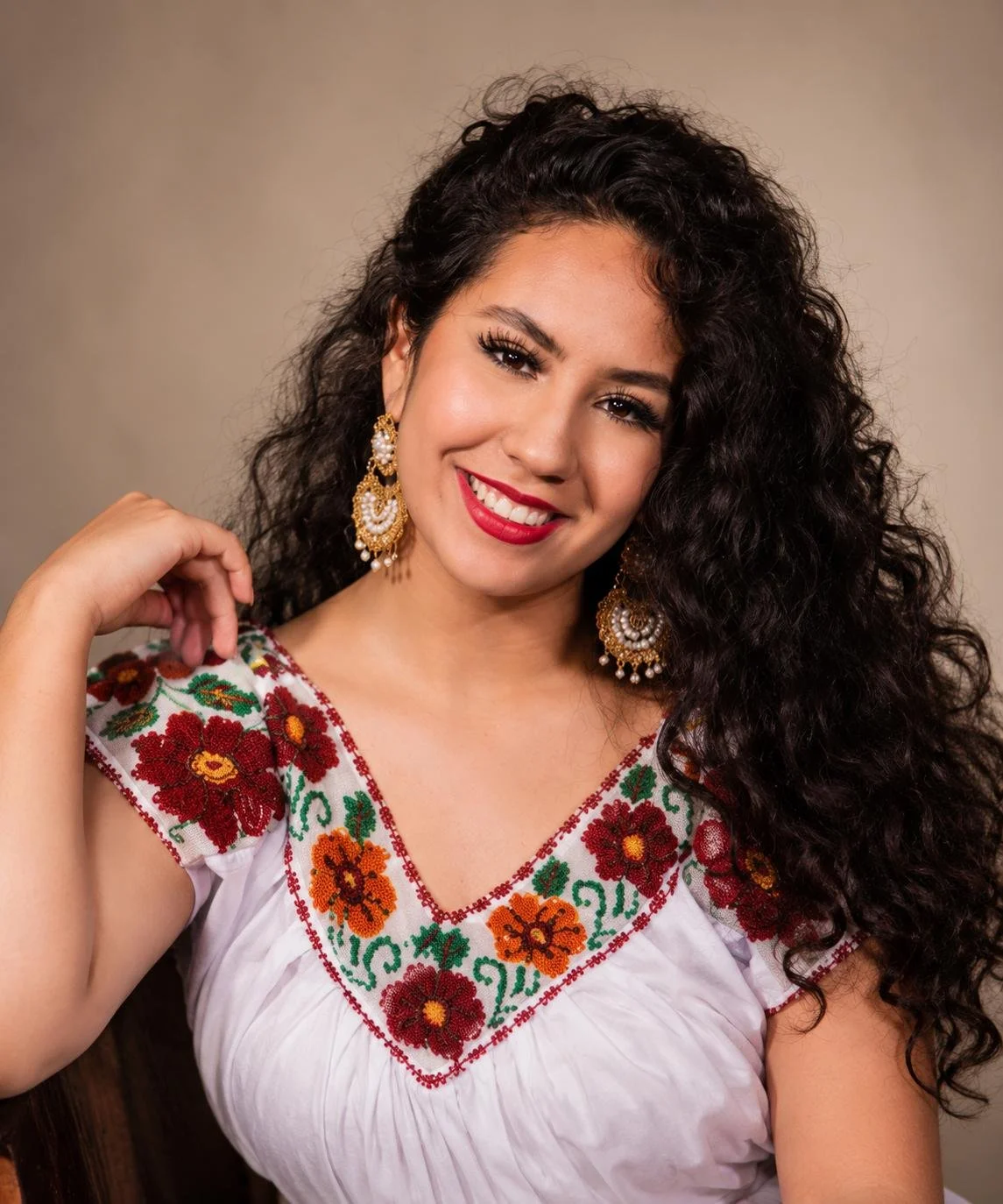 A woman with long, curly black hair, wearing traditional embroidered white blouse with colorful floral designs, and large gold earrings with pearl details, smiling at the camera.