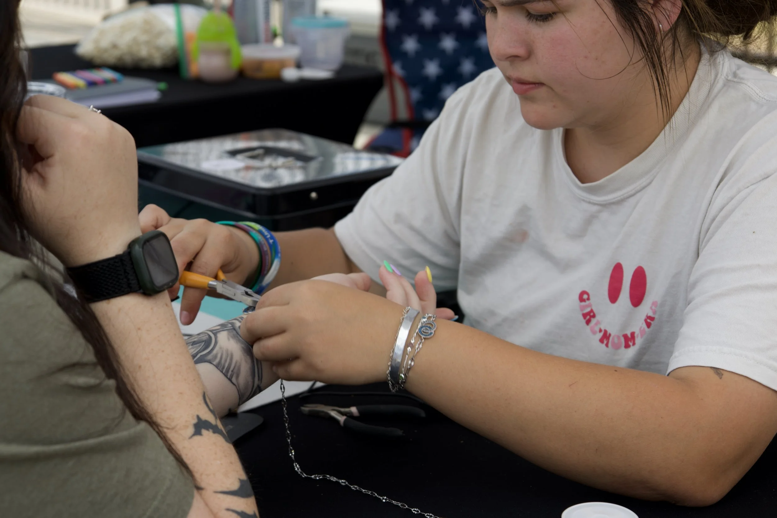 A young woman with freckles and brown hair, wearing a white t-shirt and colorful bracelets, is drawing blood from another person at a table. The person donating blood has visible tattoos on her arm and is wearing a watch. The background has various supplies and a black tablecloth.