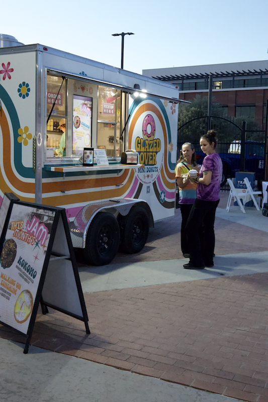 People ordering from a colorful Glazed Over mini donut food truck at an outdoor event in the evening.