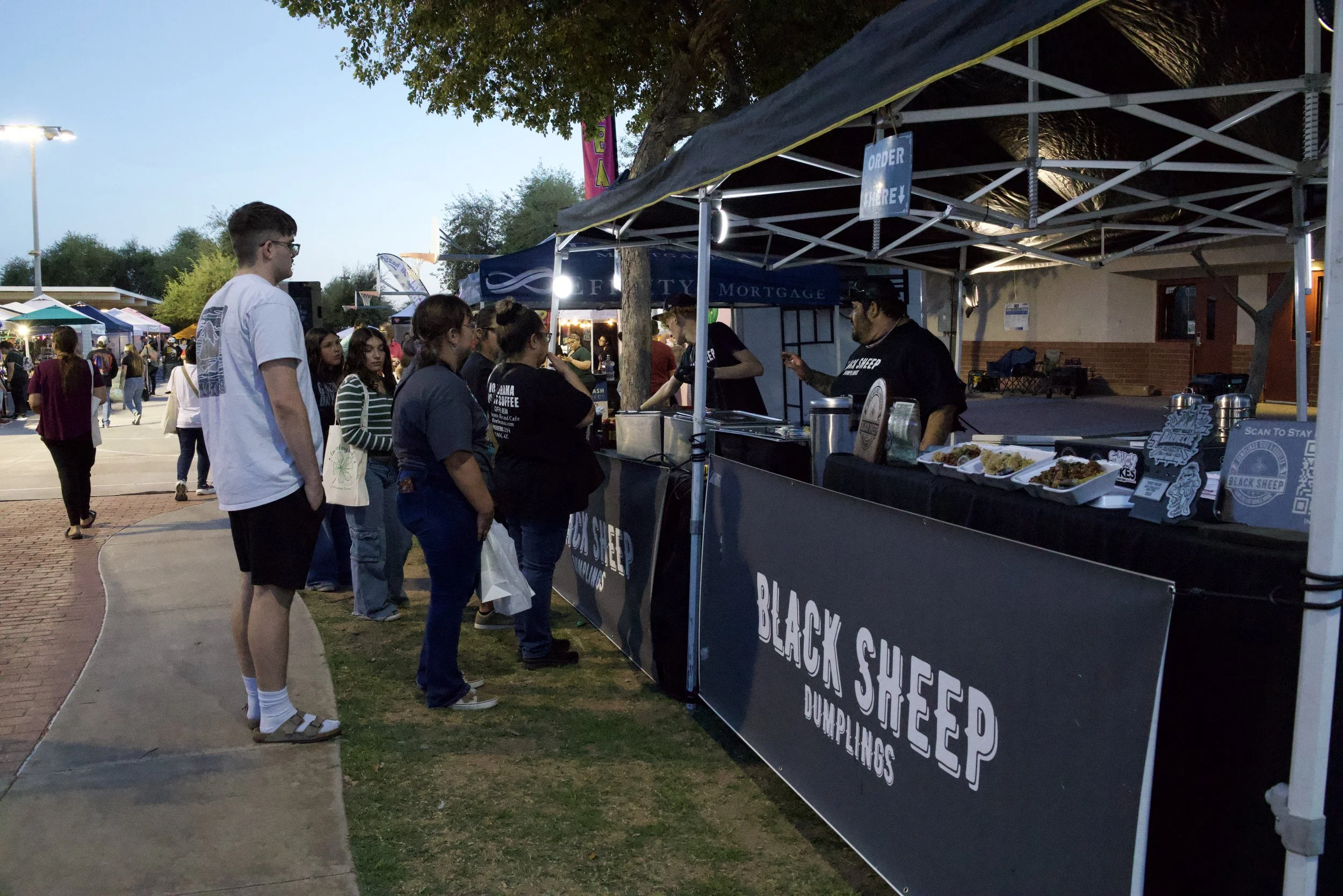 People in line at a food stand offering Black Sheep donuts and offerings at an outdoor event during dusk, with tents and trees in the background.