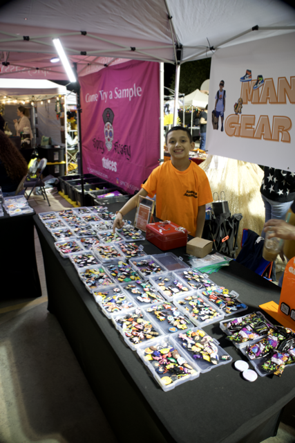 A boy in an orange shirt standing behind a table filled with colorful toys or collectibles at an outdoor market stall.