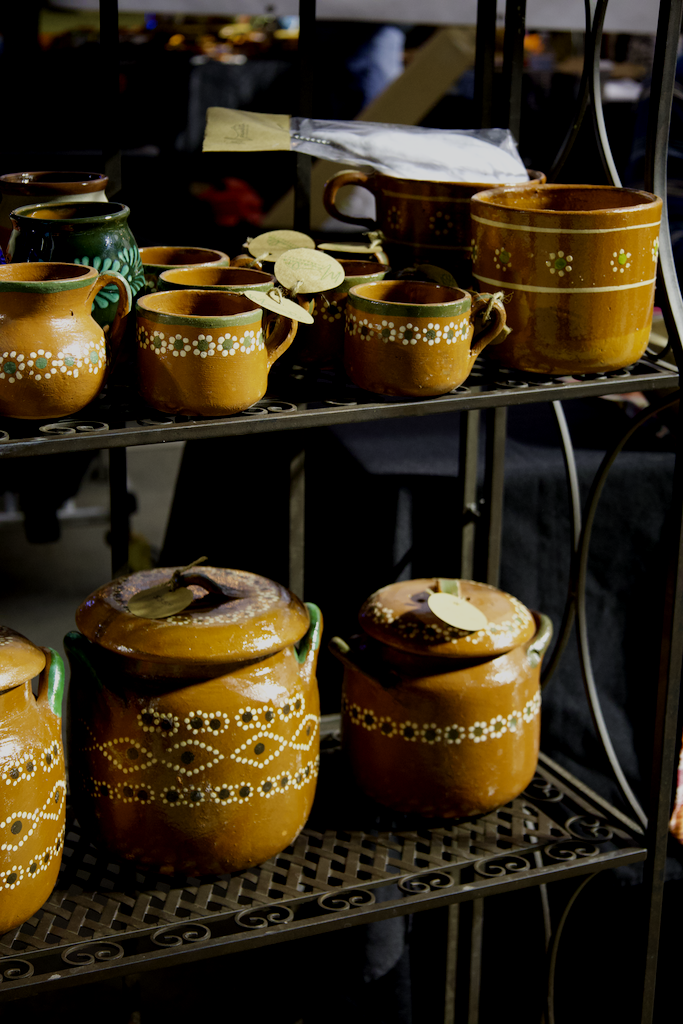 Clay pottery mugs and jars with painted floral patterns on metal shelves at a craft market.