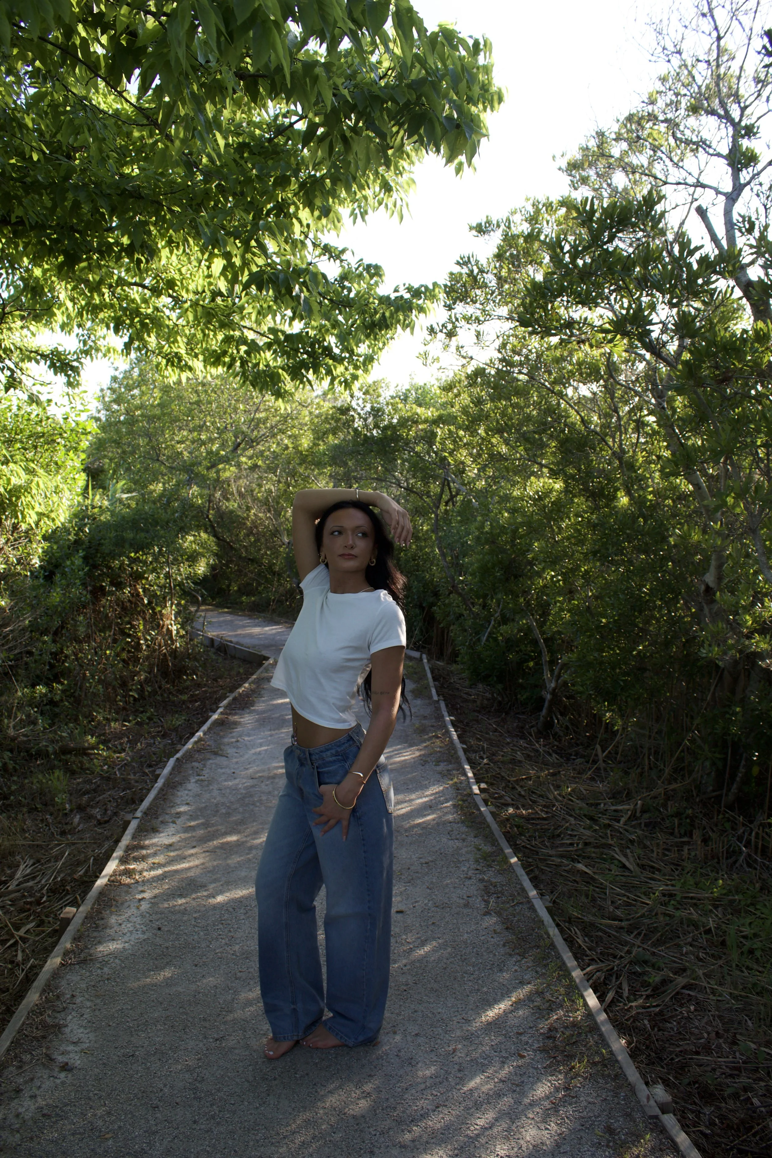 A woman with long dark hair wearing a white t-shirt and baggy jeans standing barefoot on an overgrown footpath surrounded by green trees and bushes, with her right arm raised above her head and her left hand resting on her hip, sunlight filtering through the leaves.