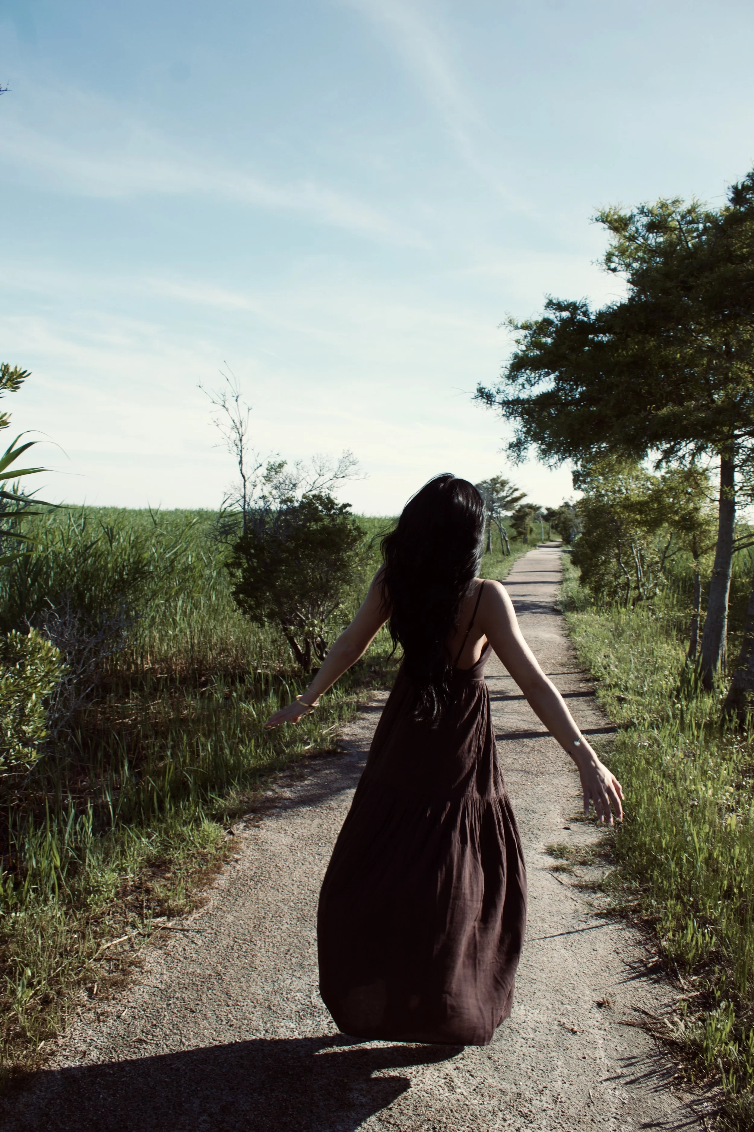 A woman in a brown dress is walking on a dirt path through tall grass and trees on a sunny day with a clear blue sky.