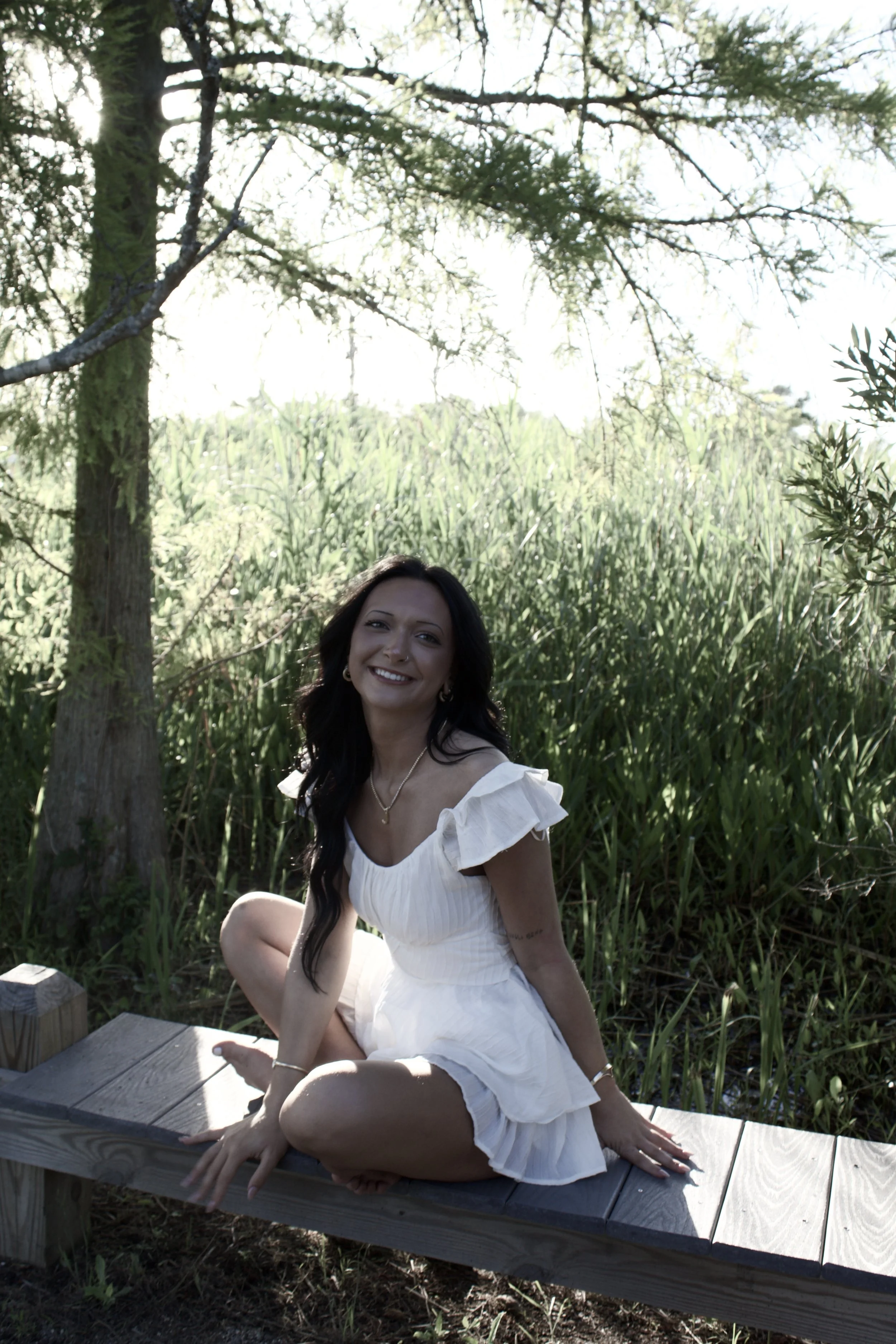 A woman with long dark hair, wearing a white dress, is sitting on a wooden bench outdoors surrounded by tall grass and trees, smiling and looking at the camera.
