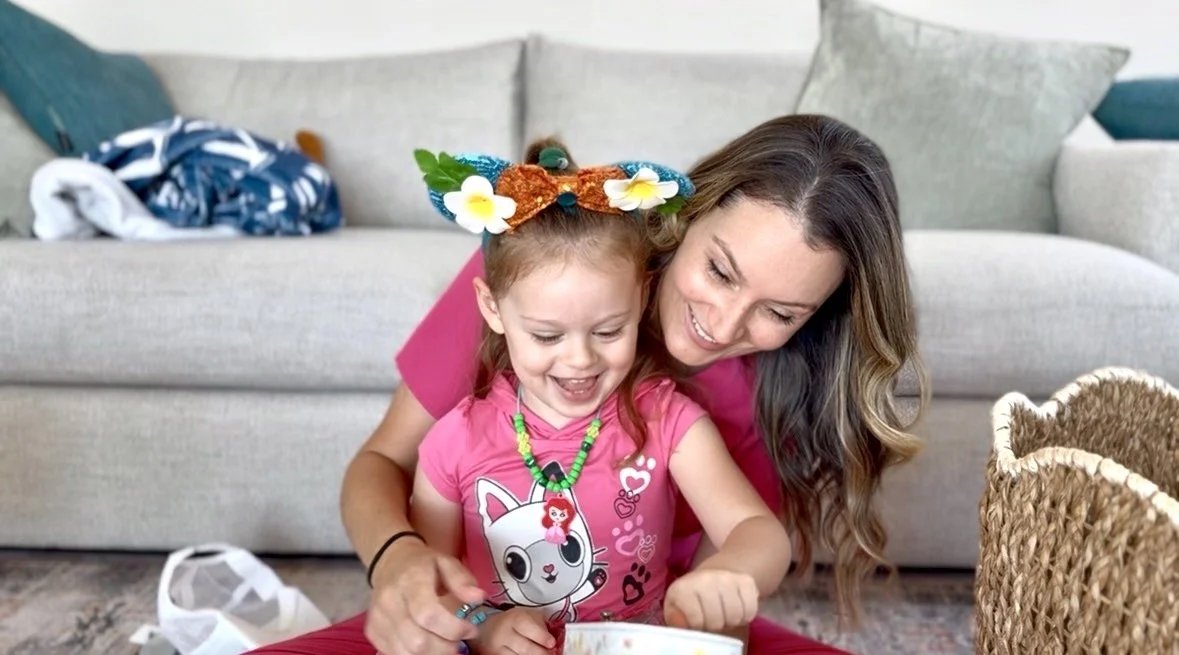 A woman and a young girl sitting on the floor in a living room, smiling and looking together. The girl is wearing a pink shirt with a cartoon character and colorful jewelry, and has a large hair accessory shaped like a butterfly. The woman is wearing a pink shirt and has long, wavy brown hair.