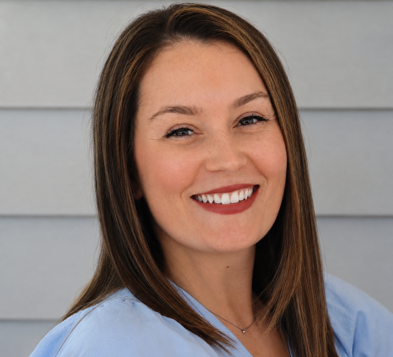 A woman with straight brown hair and a big smile, wearing a light blue top, standing in front of gray horizontal siding.
