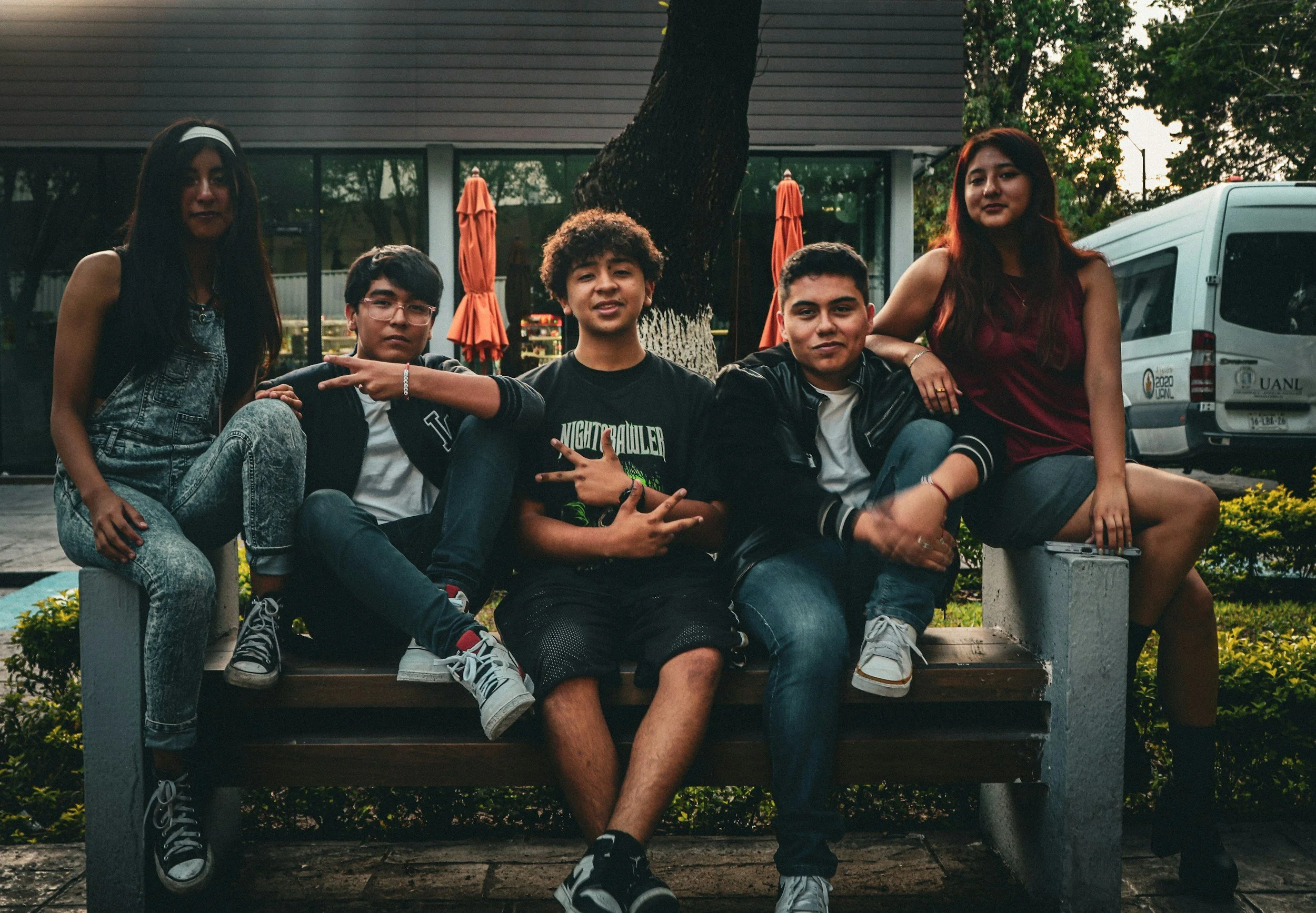 A group of five young people sitting and standing on a park bench outdoors during the evening, with trees and a building in the background.
