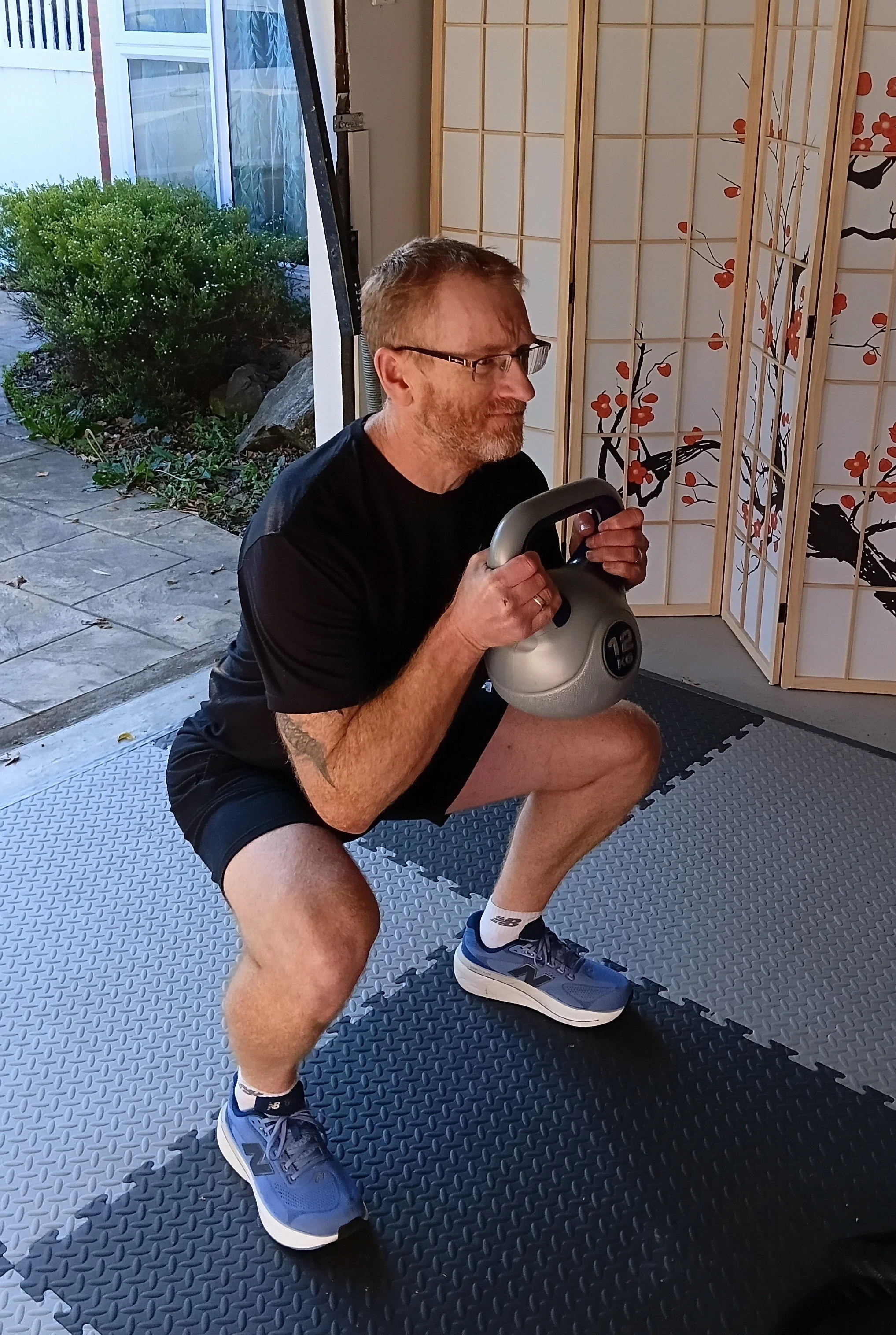 A man with glasses and a beard doing squats with a kettlebell in a gym or home workout space.