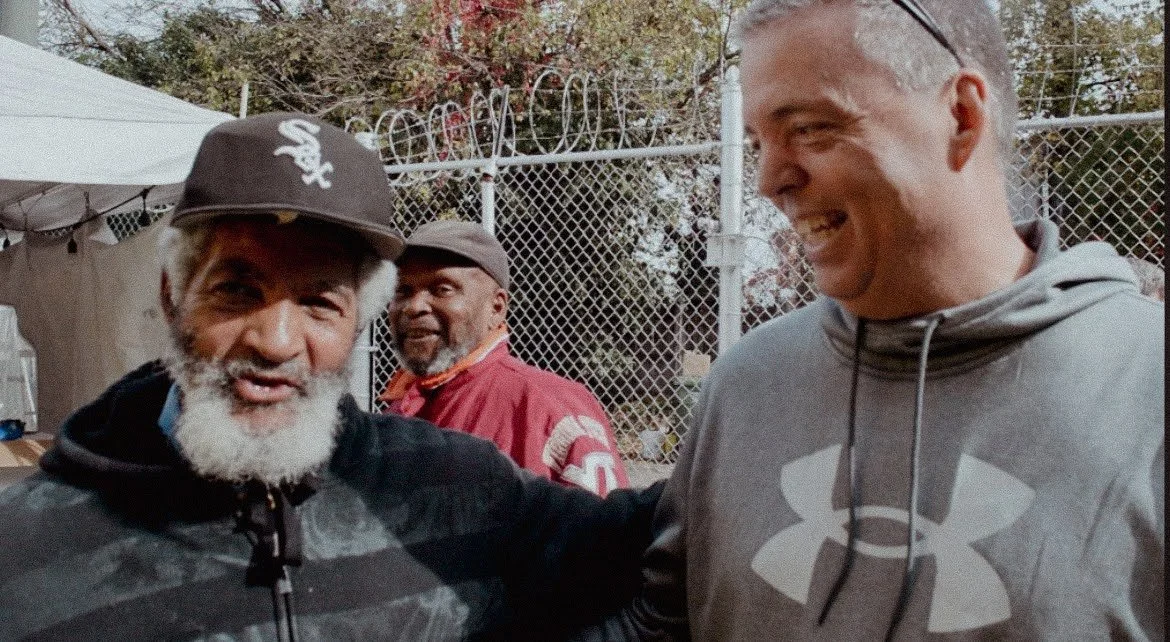 Three men smiling outdoors near a chain-link fence, one wearing a Chicago White Sox cap, another in a red jacket, and the third in a gray hoodie with a logo.