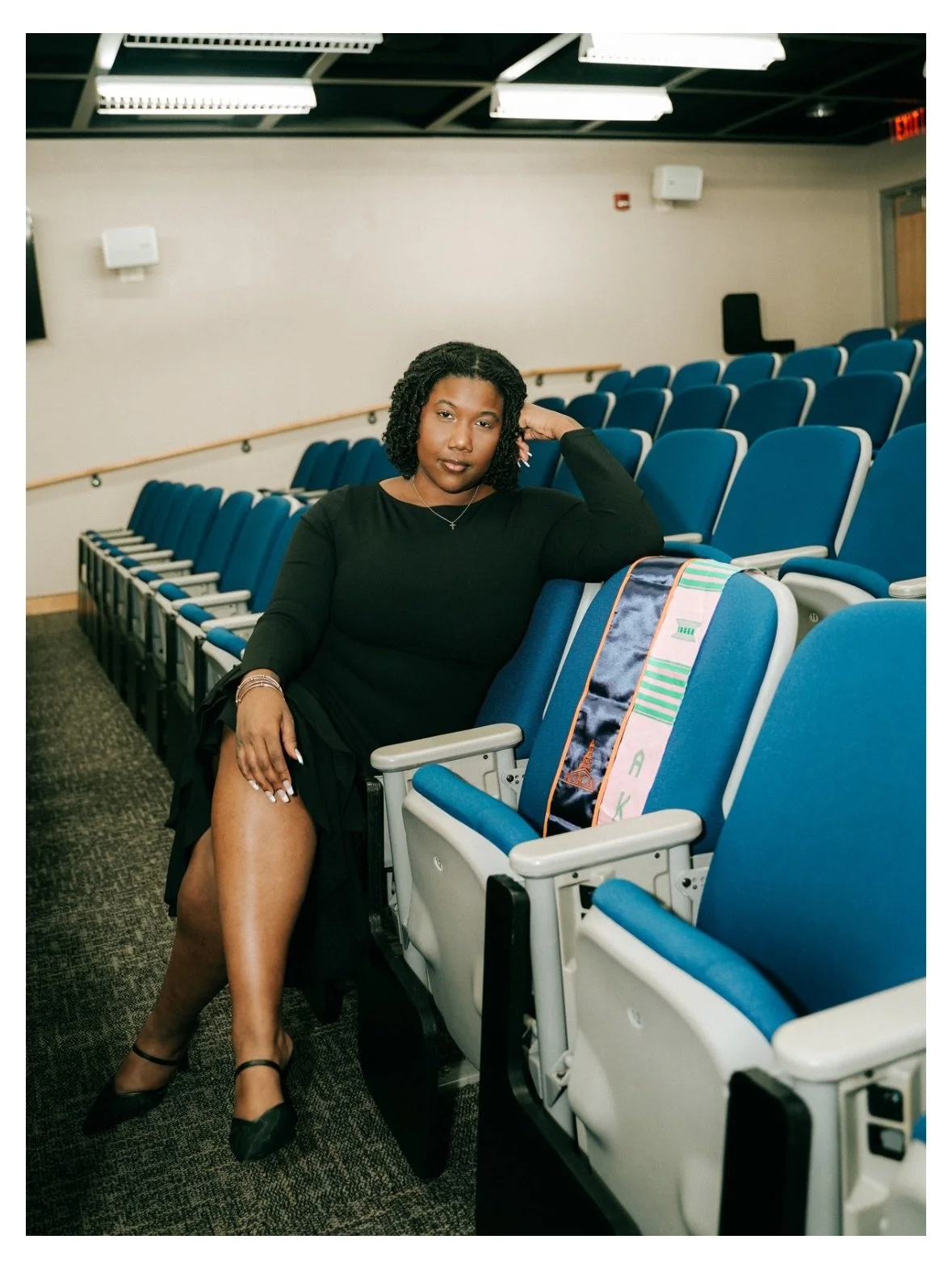 A woman sitting in a nearly empty auditorium with blue seats, wearing a black dress, black heels, and jewelry, with a colorful bag in the seat beside her.