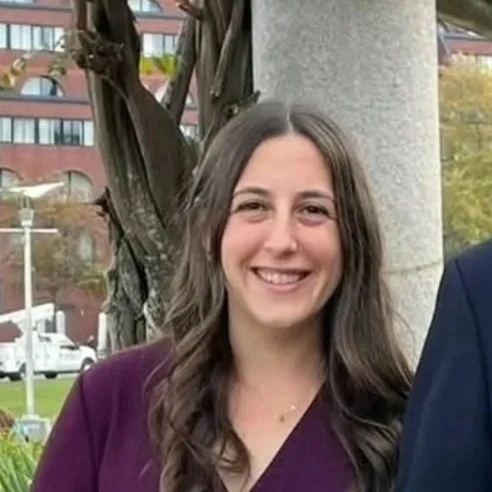A smiling woman with long brown hair standing outdoors next to a tree and concrete pillar.