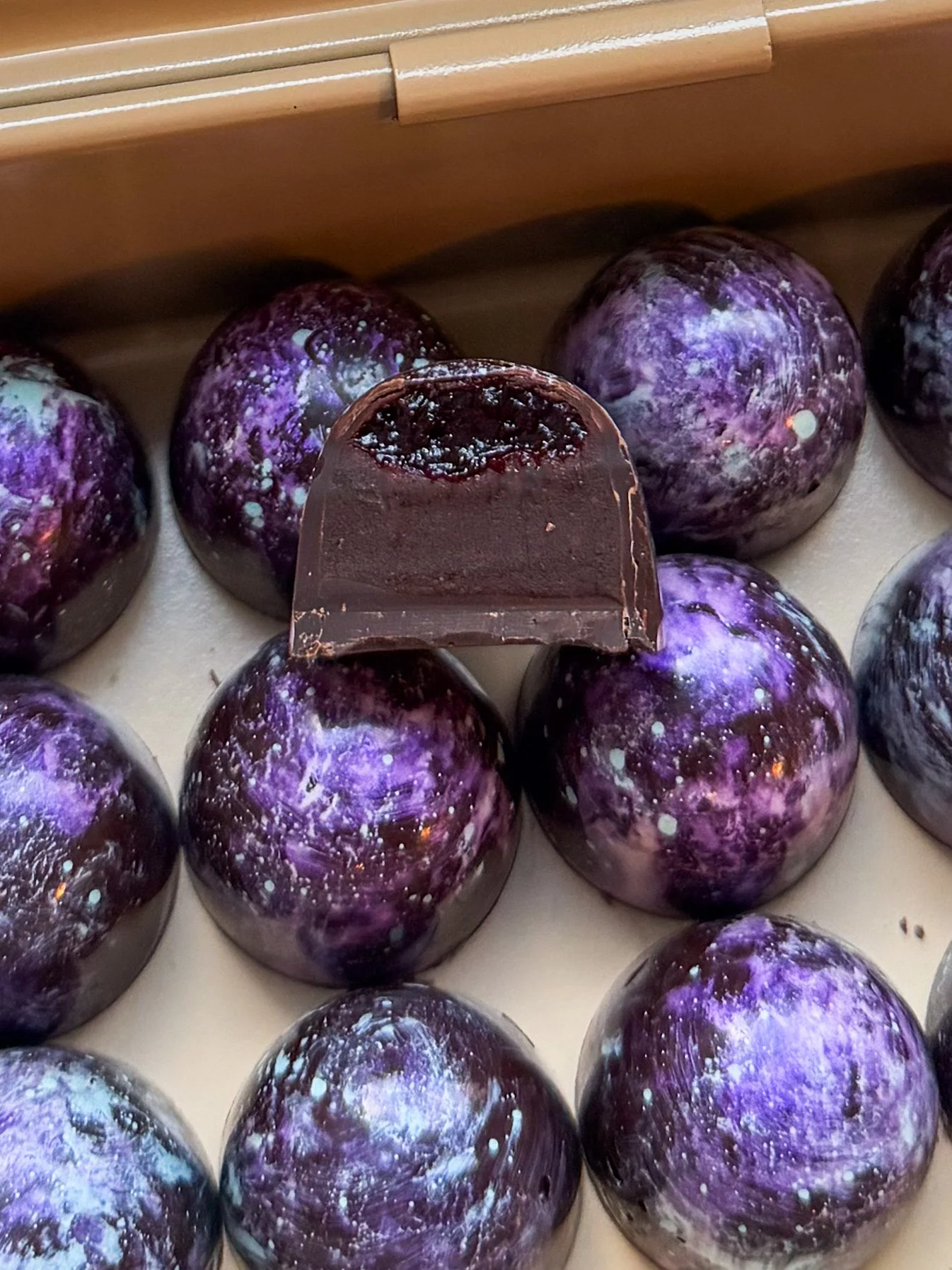 Close-up of a collection of cosmic-themed chocolates with purple, blue, and white galaxy designs, one of which is partially cut open to show a dark berry filling.