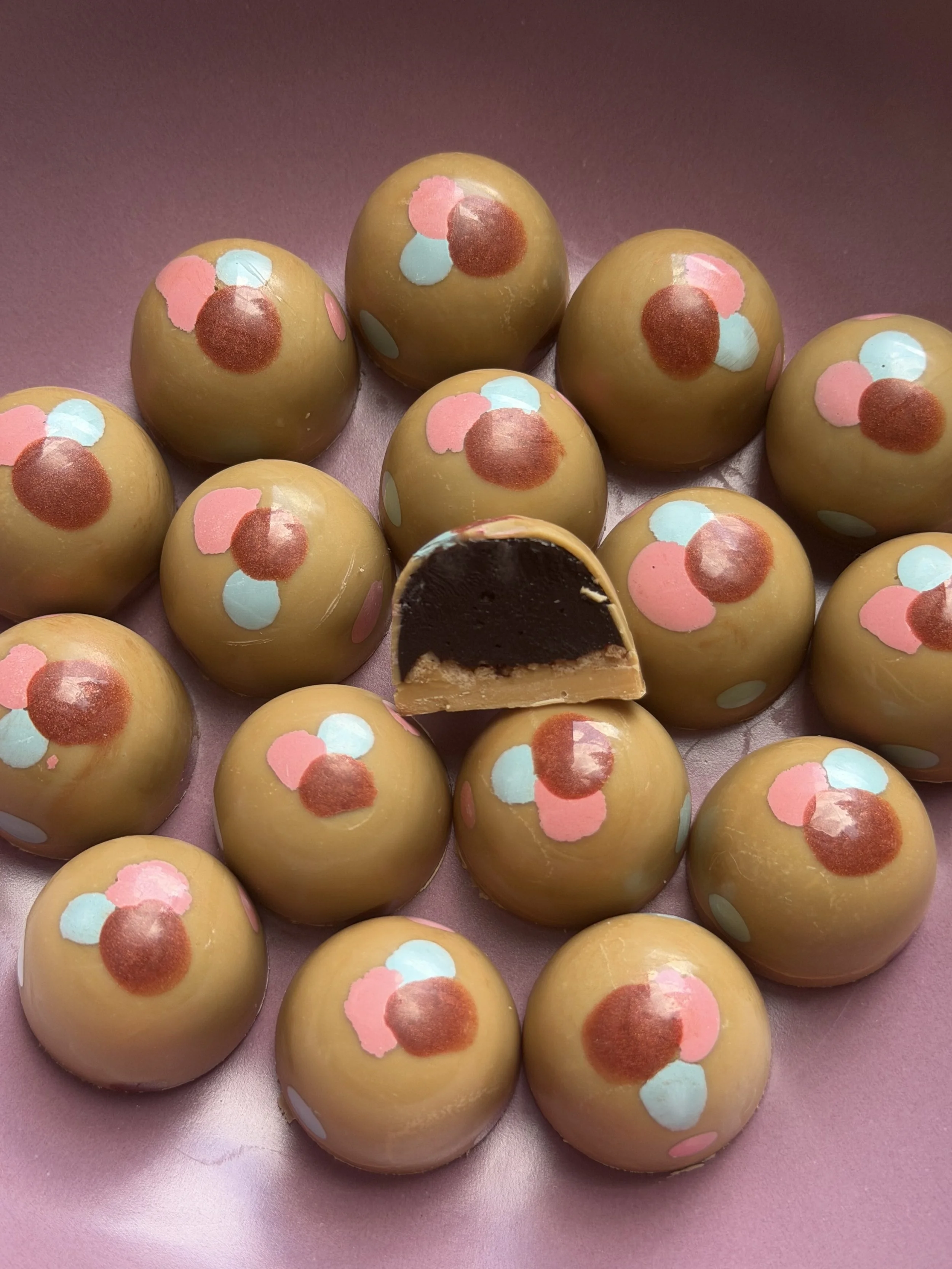 A collection of round chocolates with multicolored pastel sprinkles on top, some with a cross-section showing caramel and dark chocolate fillings inside, arranged on a pink surface.