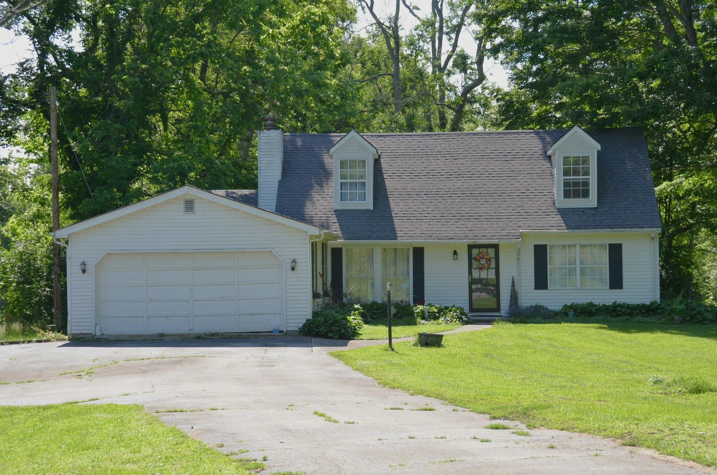 A white two-story house with black shutters and a dark gray roof, featuring a front door decorated with a wreath, visible in a lush green yard with a driveway and surrounded by tall trees.