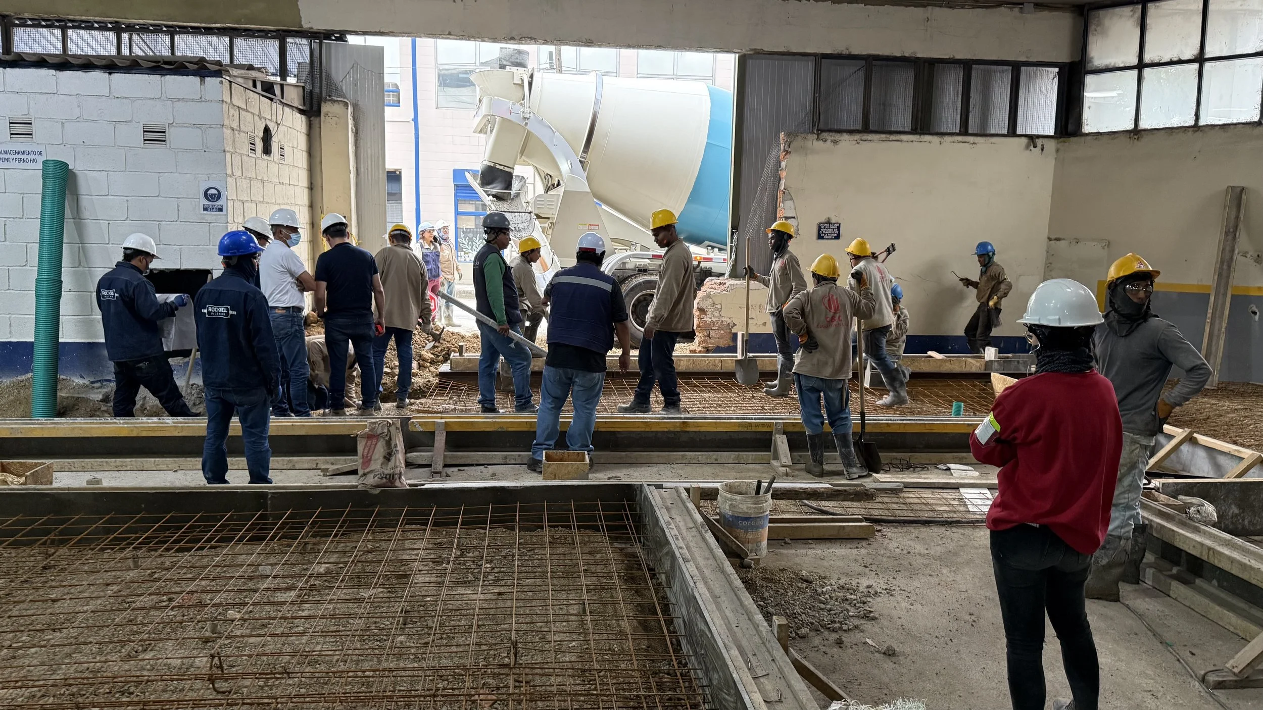 Varias personas con casco de seguridad trabajando en la construcción de un piso de concreto, con una mezcladora de cemento y varillas de refuerzo en un espacio abierto