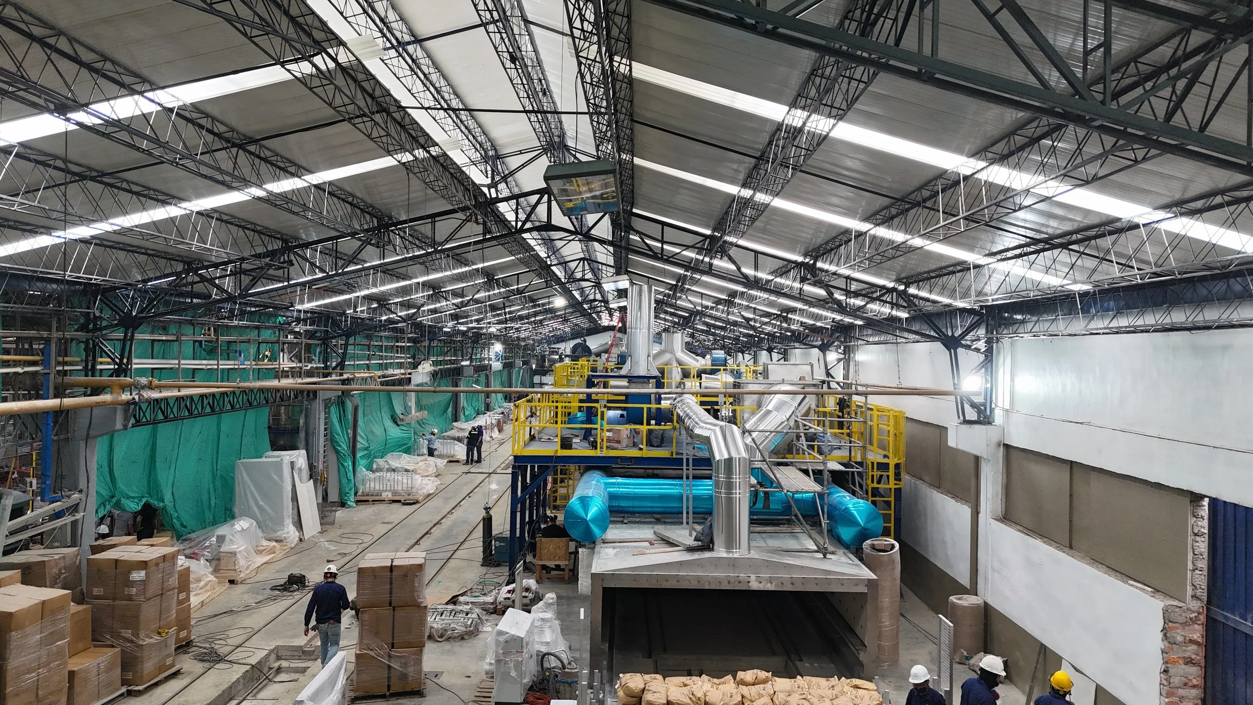 Interior de una planta industrial en construcción con estructura metálica, tuberías, cajas y personal trabajando.