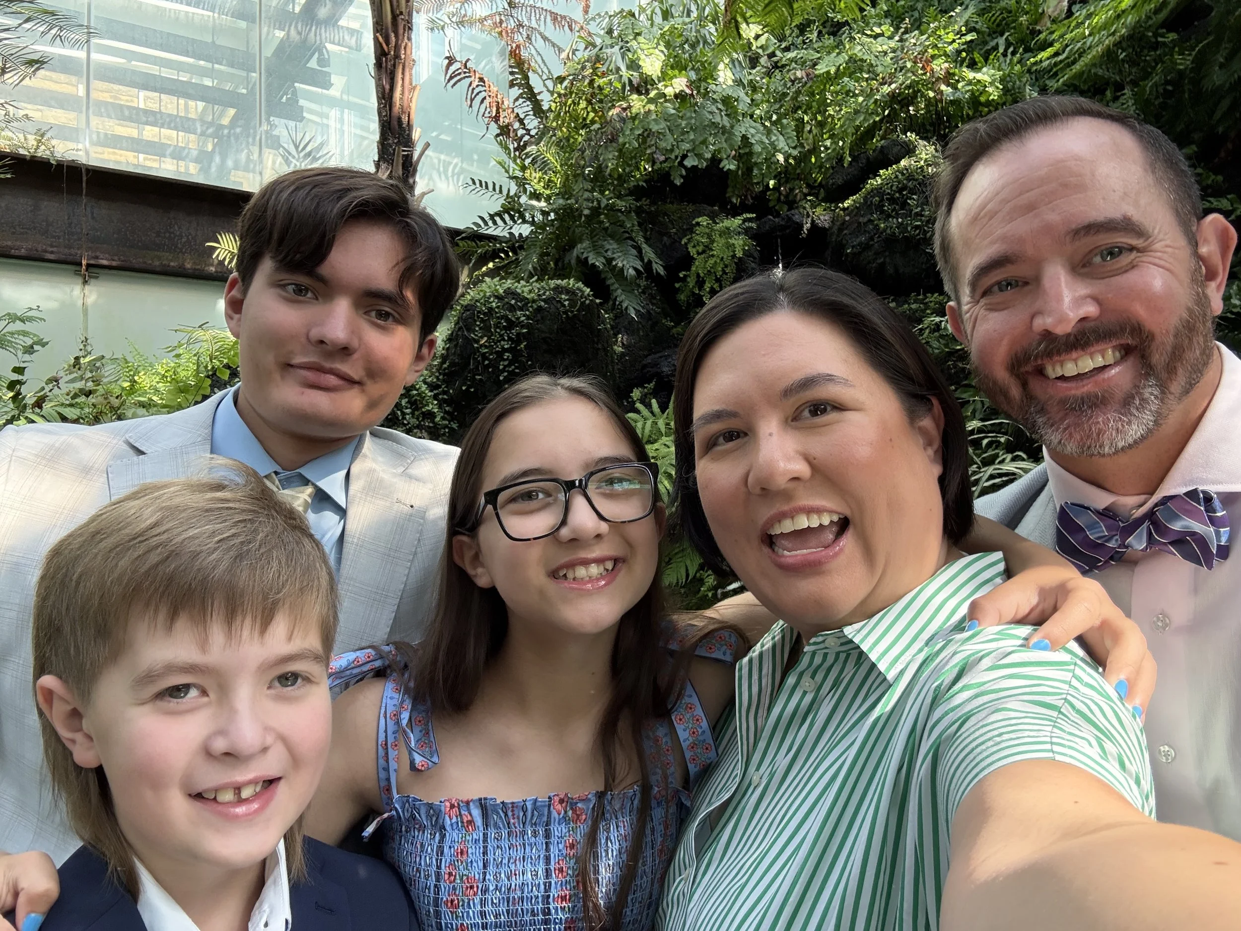 Kim Humann with her family smiling for a selfie in a greenhouse. Kim and her two sons, daughter, and husband are dressed in suits, ties and dresses with green ferns appearing in the background.