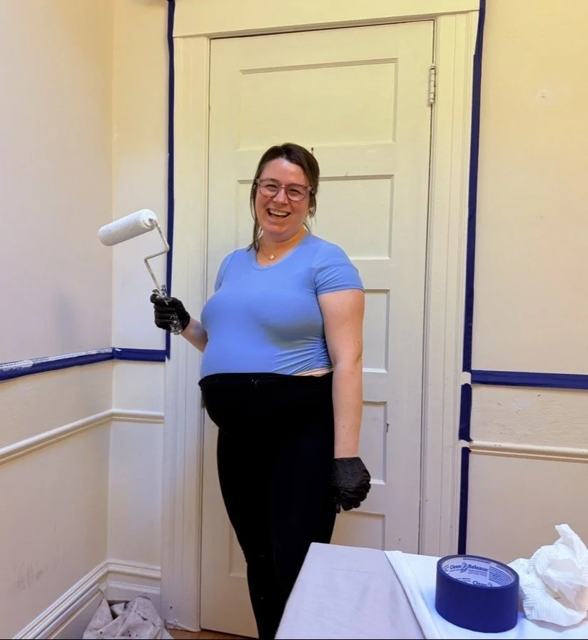 Woman smiling while painting a room with a paint roller, wearing black gloves, in a partially painted room.