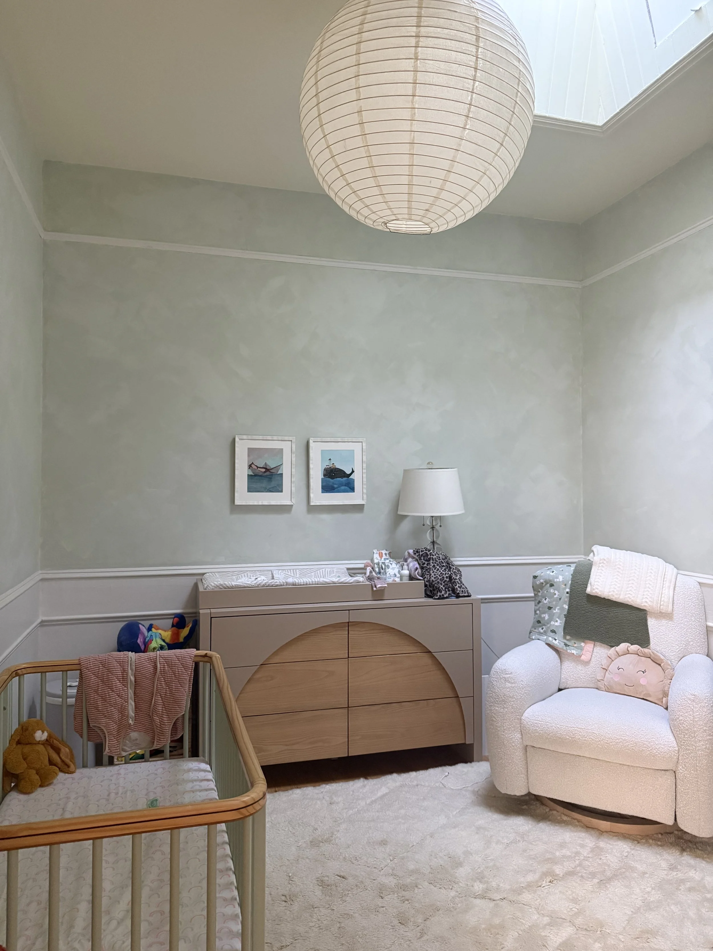 A nursery room with a white armchair, plush toys, a changing table, a crib with hanging blankets, two framed pictures on the wall, a table lamp, and a spherical paper lantern hanging from the ceiling.