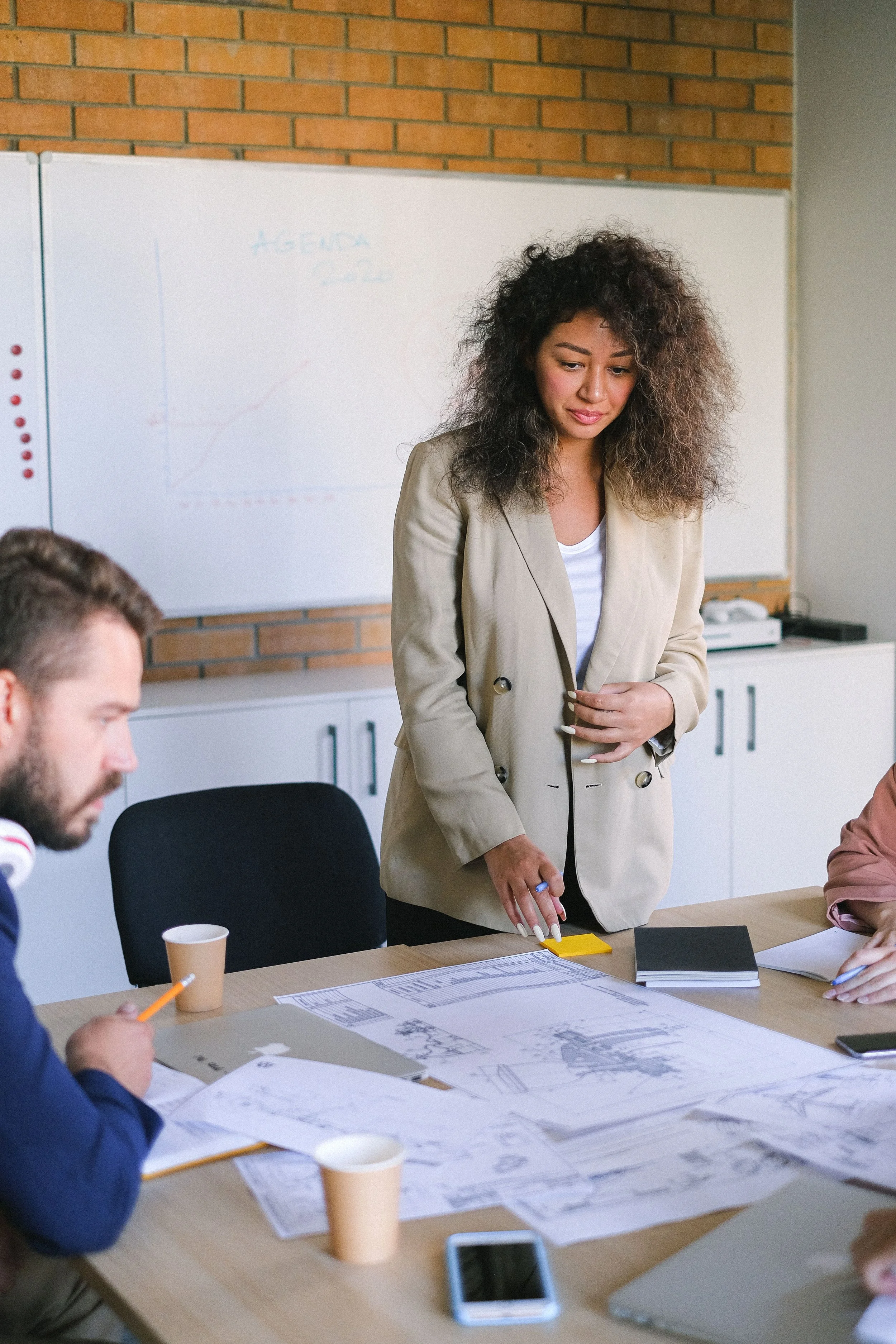 A woman with curly hair standing at a conference table with architectural plans, talking to two men. There are coffee cups, papers, and a smartphone on the table. A whiteboard in the background has a graph and writing.