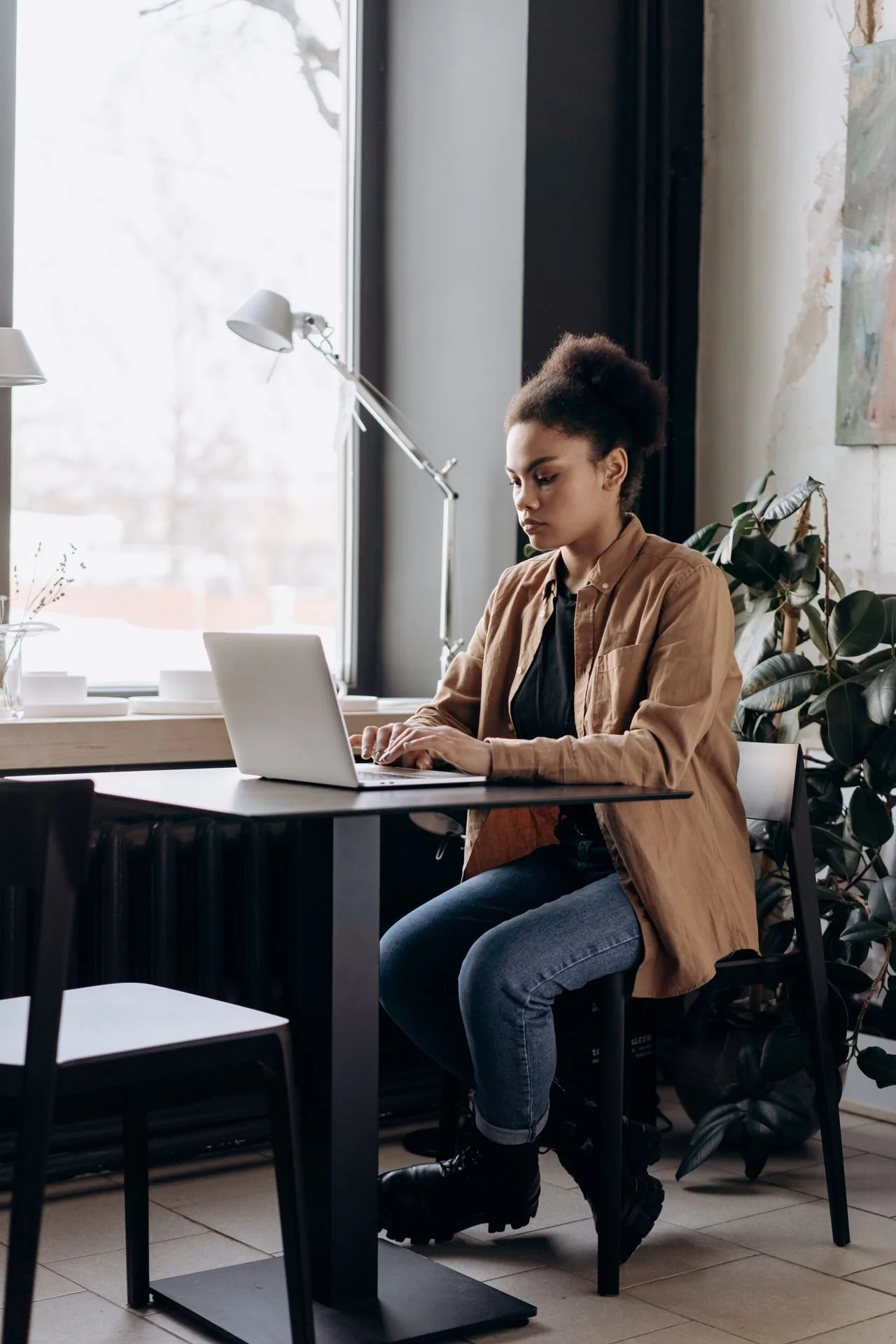 A woman with curly hair, wearing a tan jacket, sits at a black table working on a silver laptop in a bright, modern room with large windows and indoor plants.