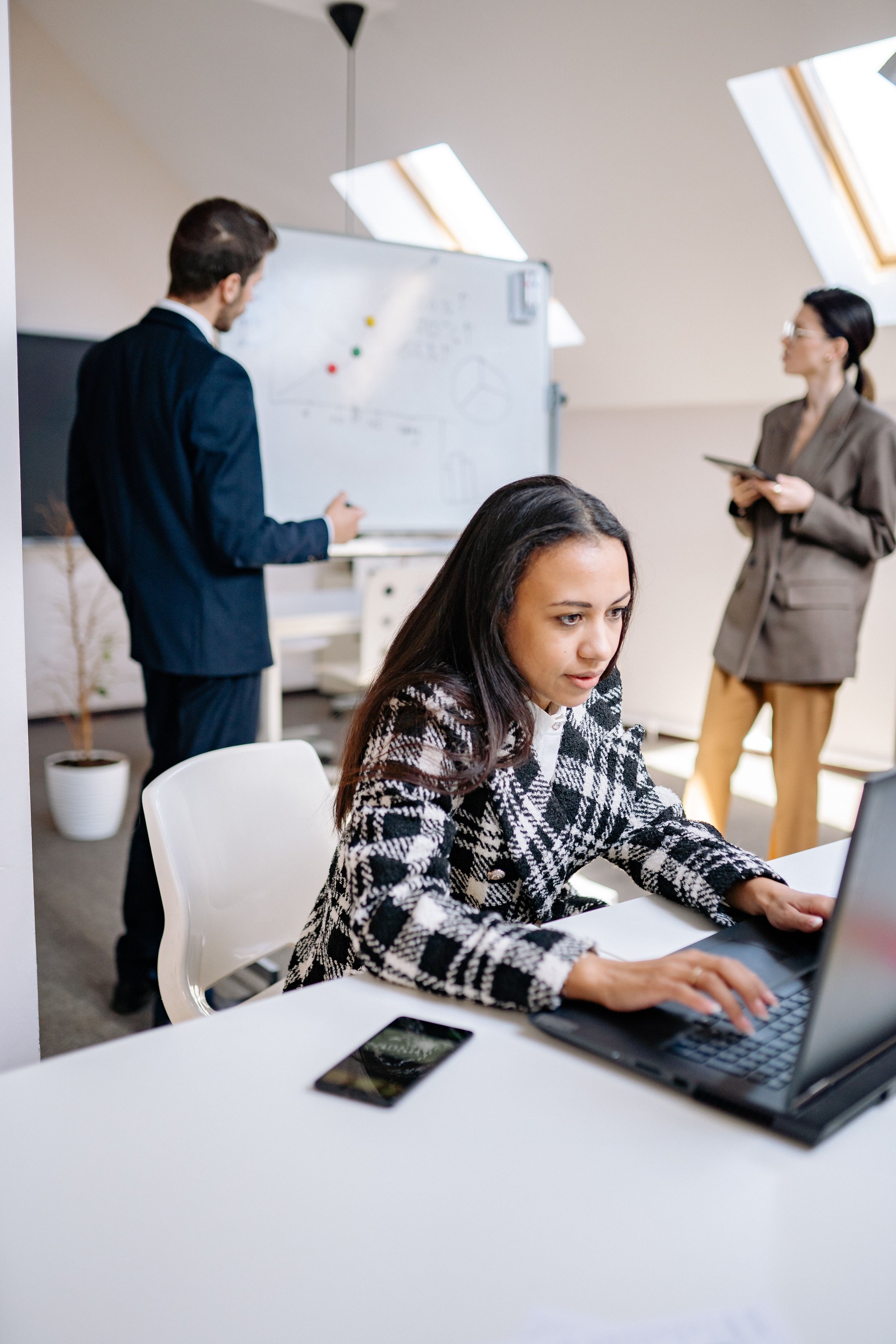 A woman with long dark hair sitting at a white desk, using a laptop. A smartphone is on the desk nearby. In the background, two people are standing, one man in a dark suit and a woman in a brown jacket, discussing near a whiteboard.